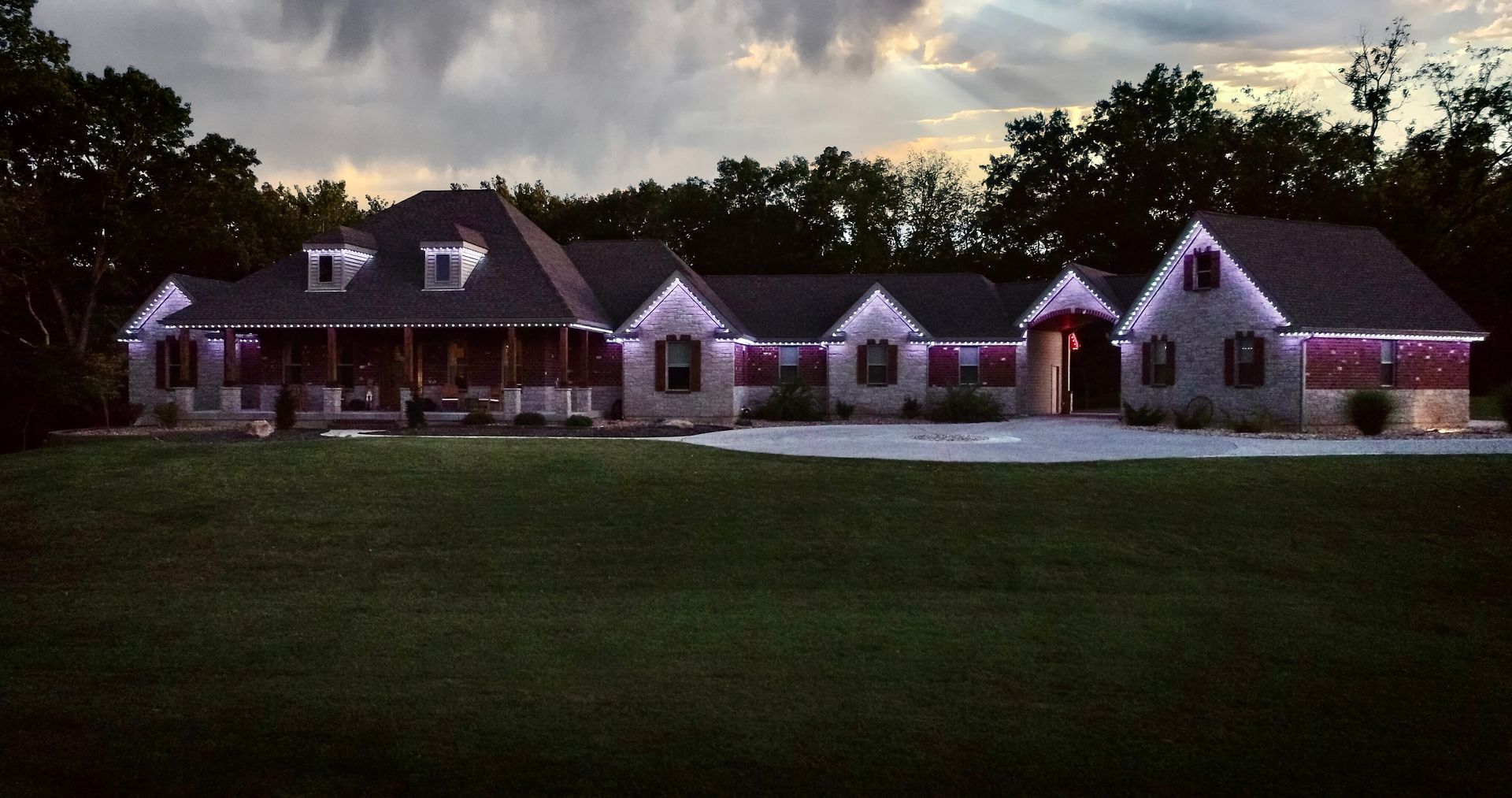 Large stone house with lit windows at dusk, set behind a wide green lawn under a cloudy sky