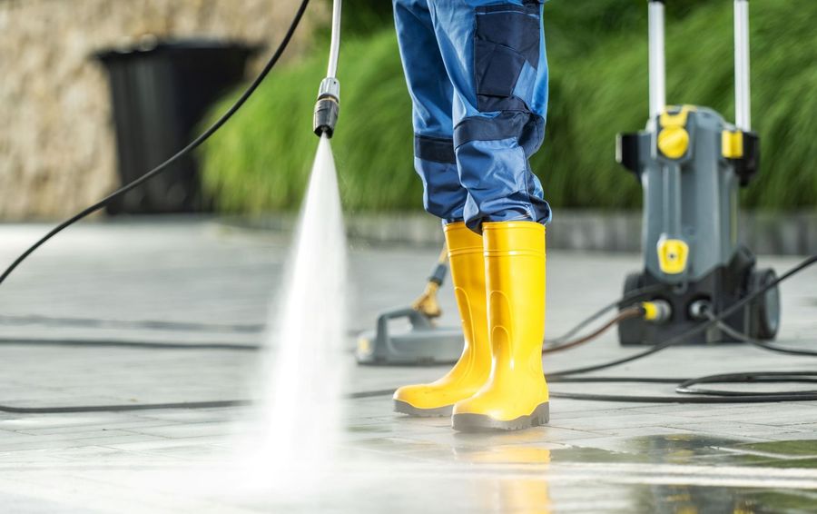 Person in yellow boots pressure-washes a wet concrete surface outdoors.