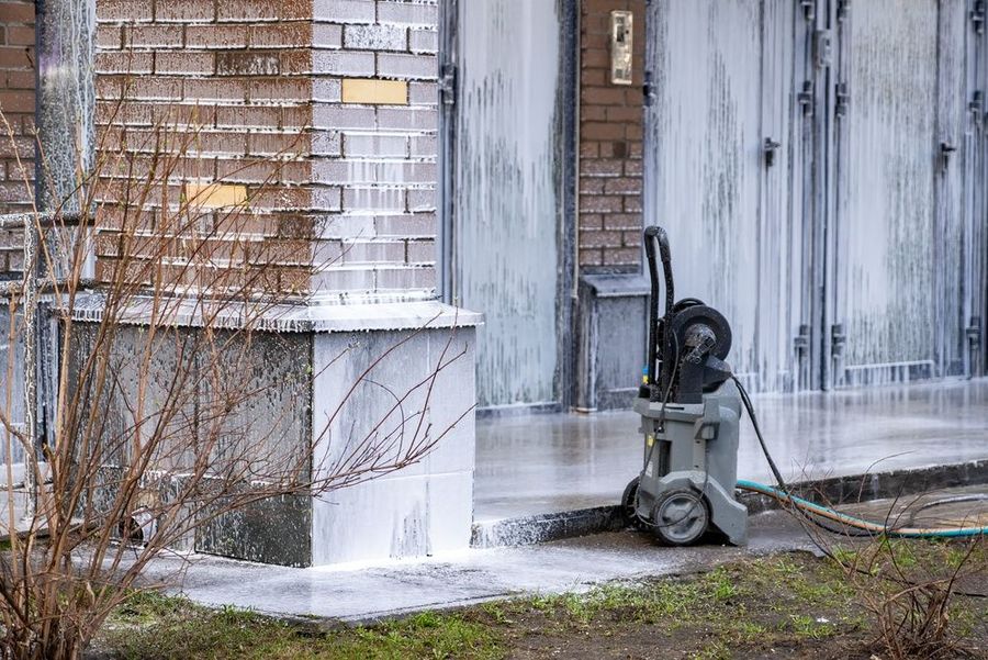 Pressure washer cleaning a brick house exterior near a door and window, with water spraying across the wall.