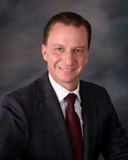 Smiling man in a dark suit and red tie against a gray studio backdrop