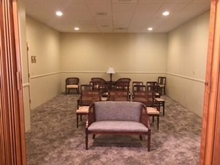 Small waiting room with rows of wooden chairs, a loveseat, and a lamp against beige walls.