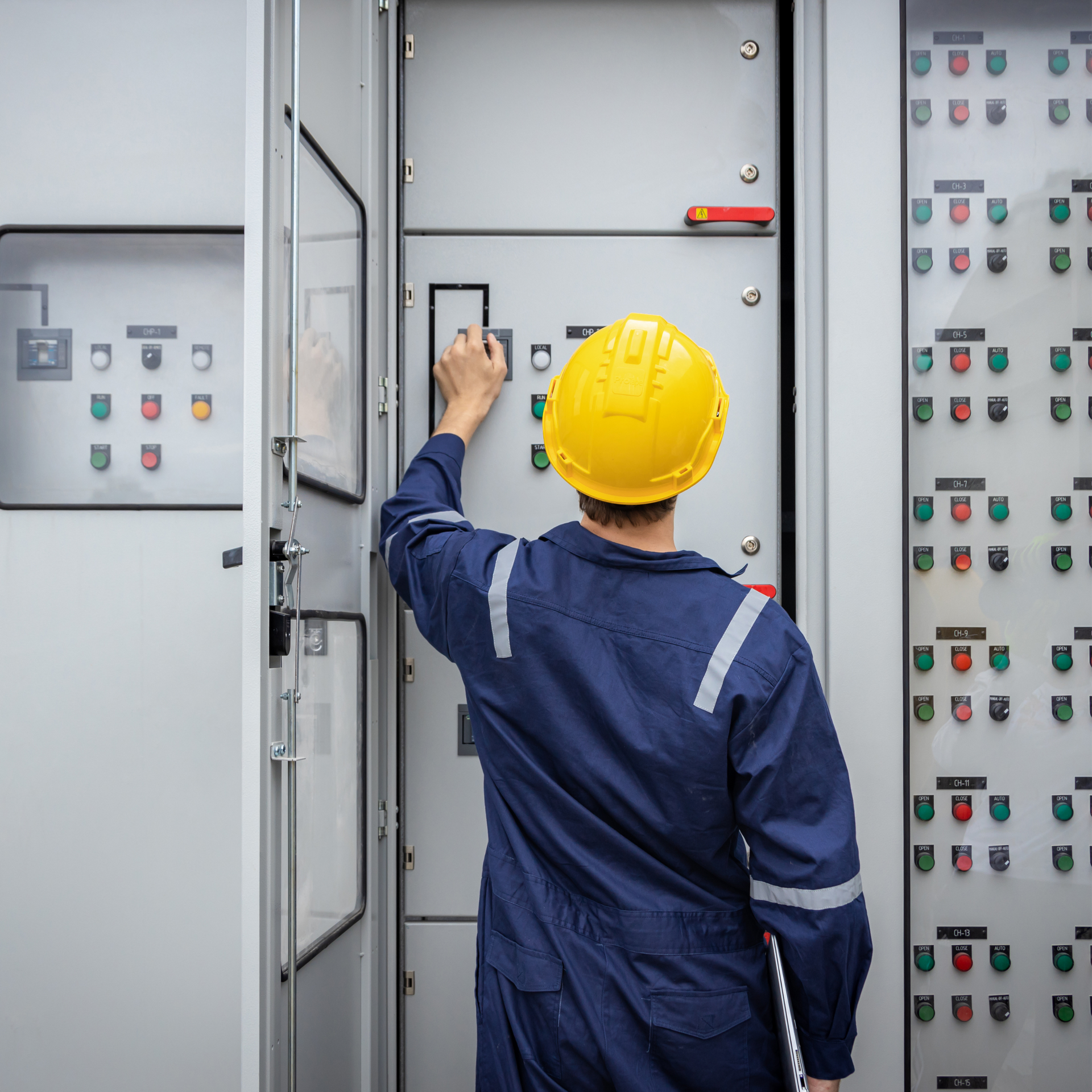 Worker in yellow hard hat inspecting an electrical control panel in an industrial room