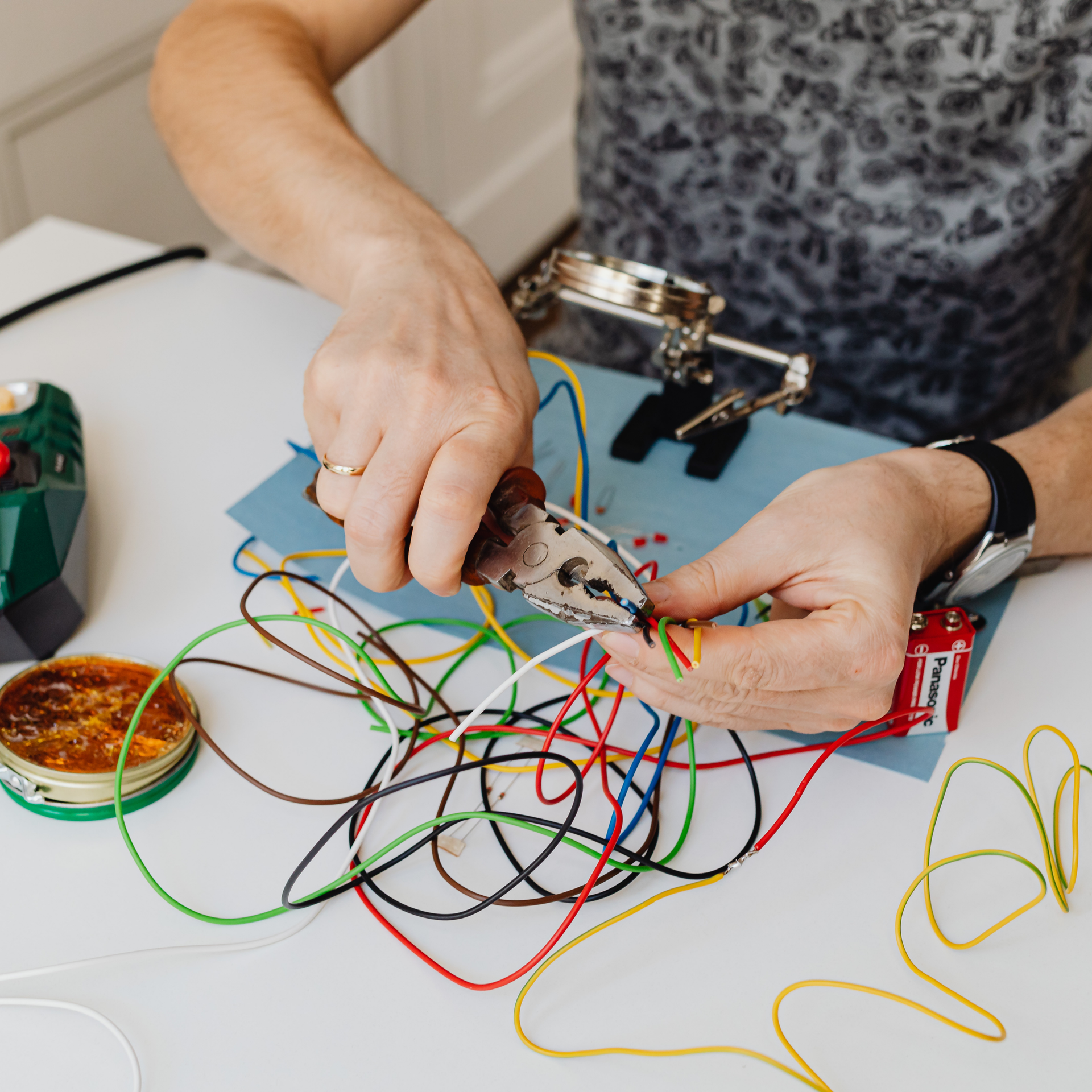 Hands using blue-handled pliers to adjust a small black component on a white cable, close-up.