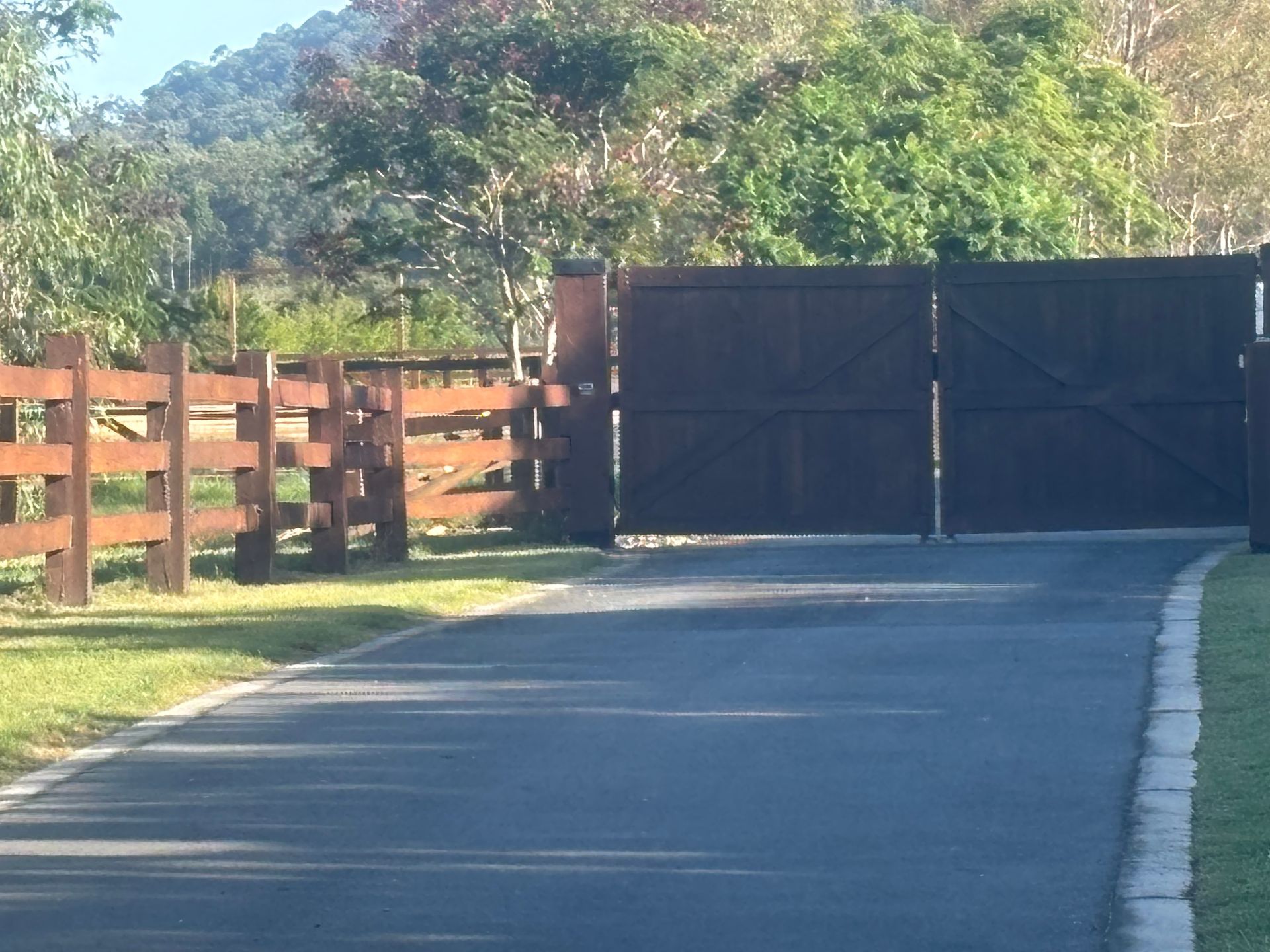 Wooden gate and fence along a paved driveway with trees in the background. — Keith Dellit Rural Fencing in Yandina Creek, QLD