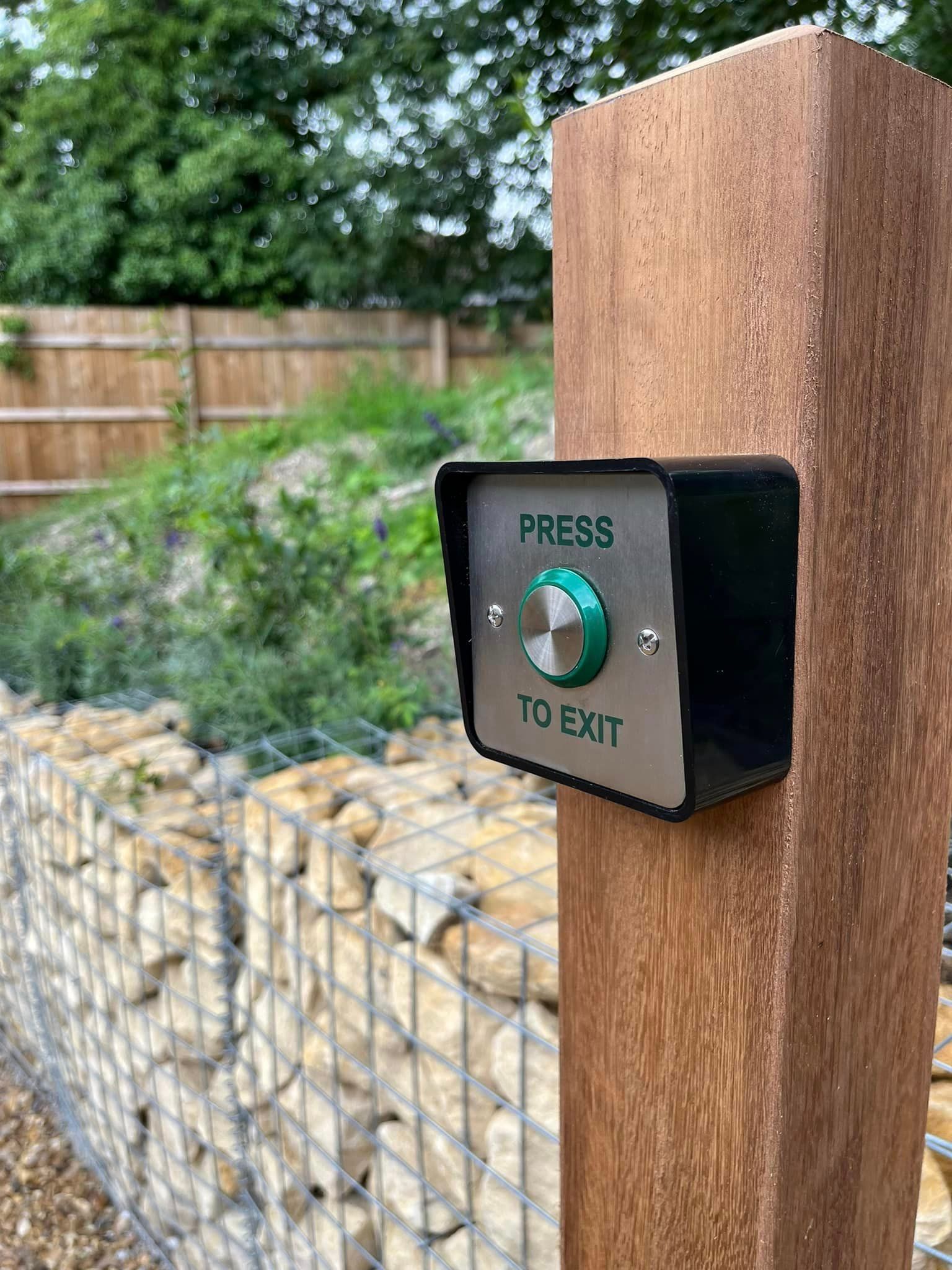 Black press-to-exit button mounted on a wooden post beside a stone wall and fence.
