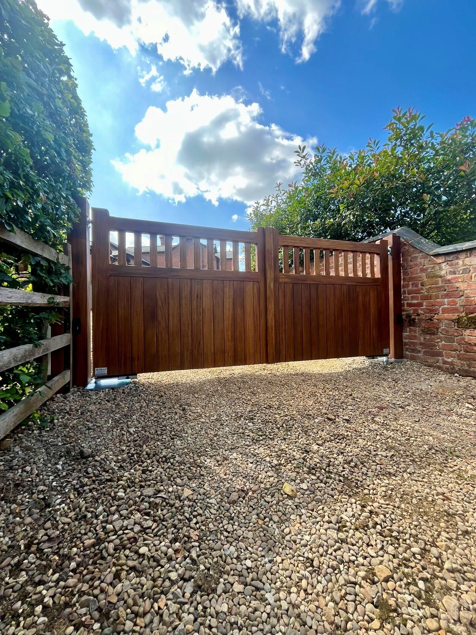 Wooden driveway electric gate with gravel foreground, brick wall, and sunny blue sky