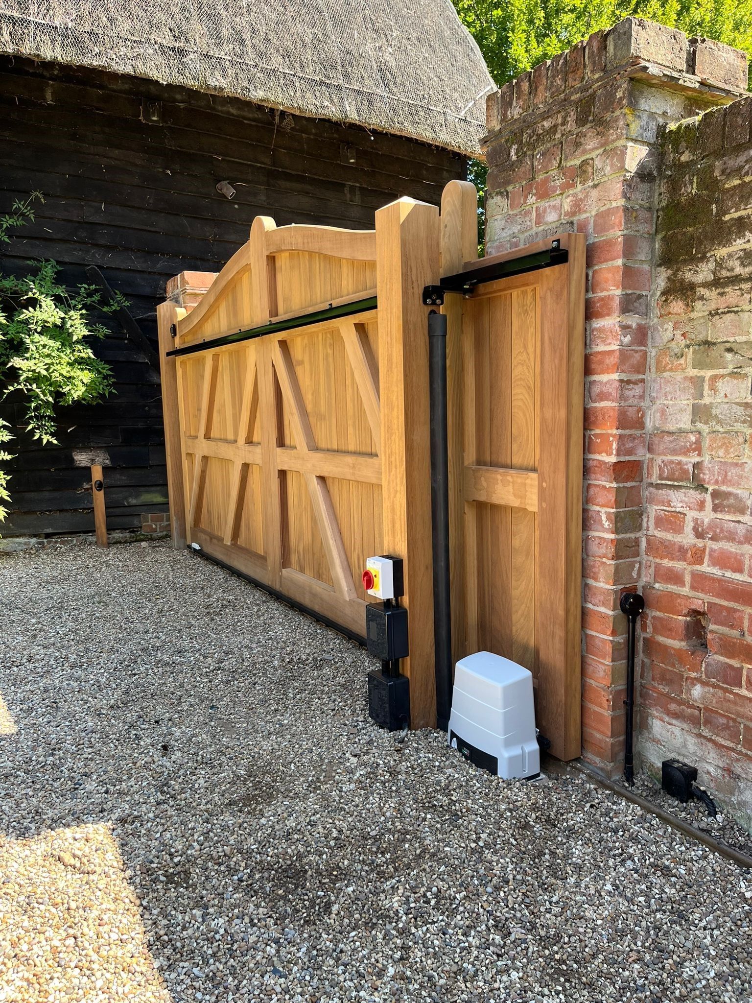Wooden driveway gate set in a brick wall beside a gravel courtyard and thatched building.