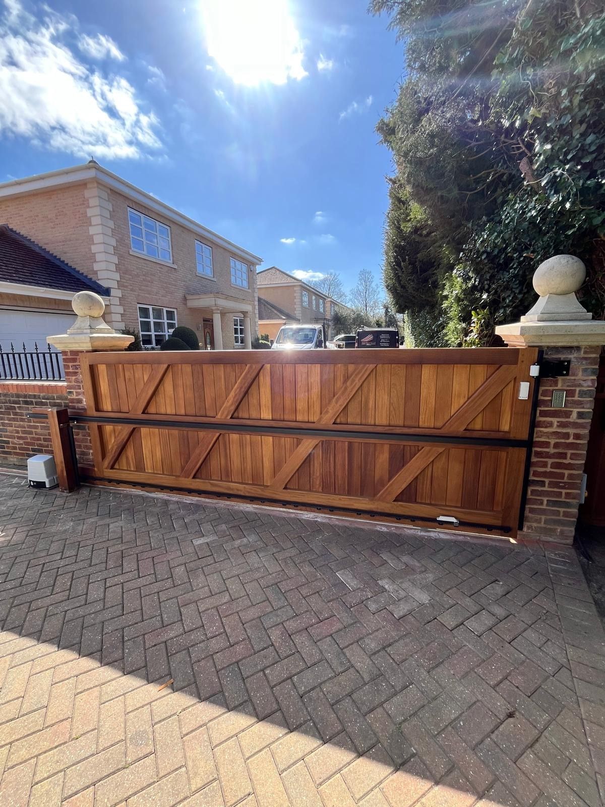 Wooden driveway gate with brick pillars in front of a house, under a bright blue sky.