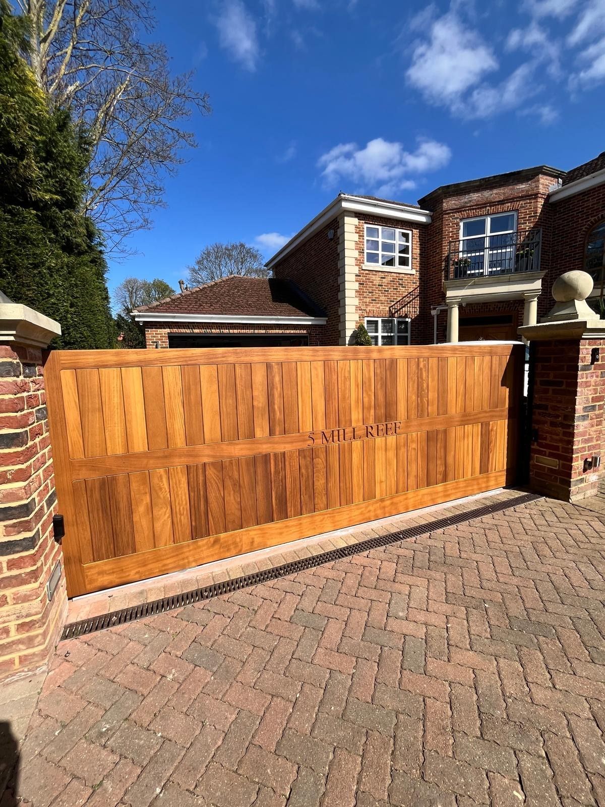 Wooden driveway gate between brick pillars in front of a brick house on a sunny day