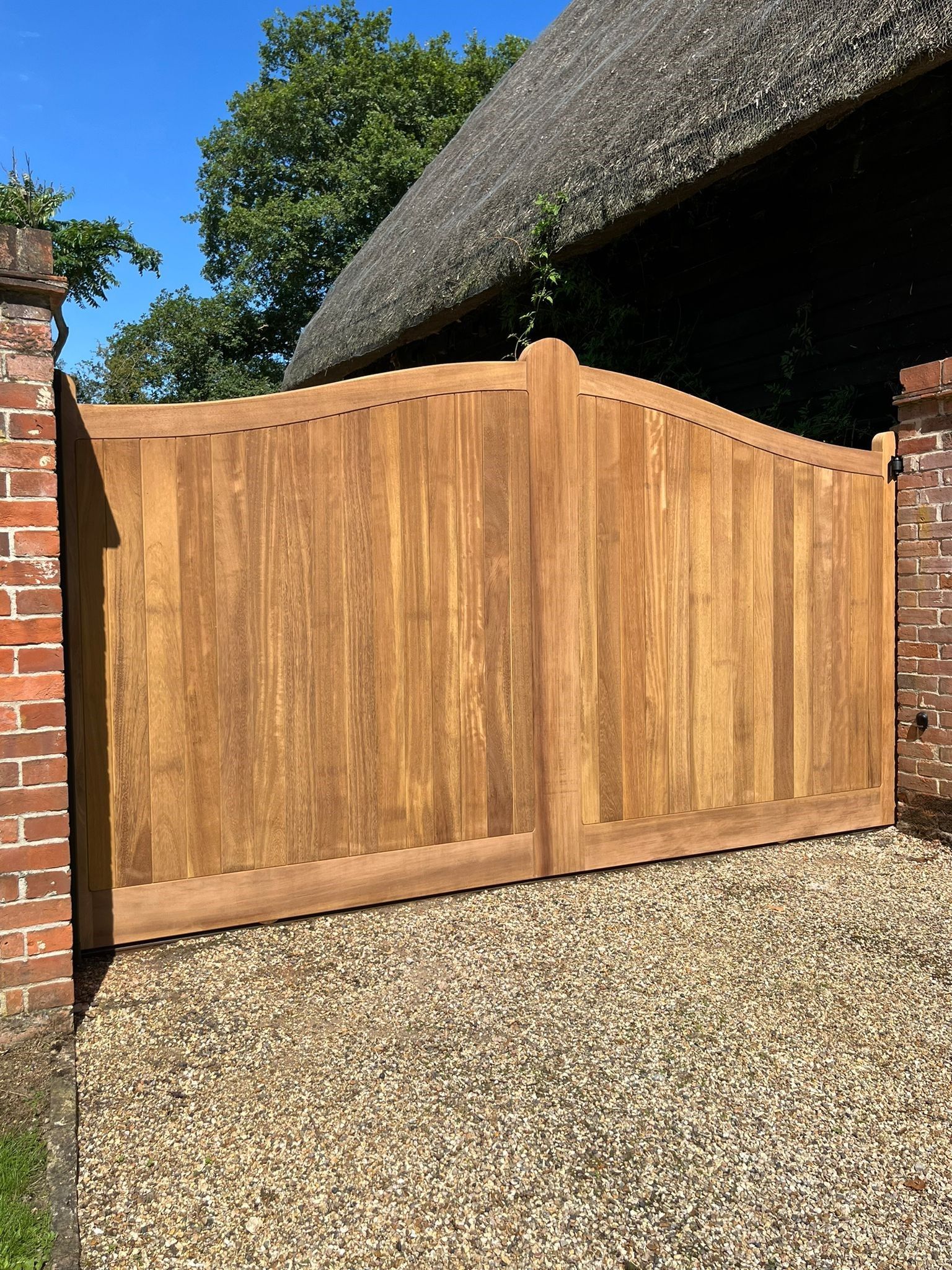 Wooden driveway gate beside a brick wall and thatched roof cottage on a gravel drive