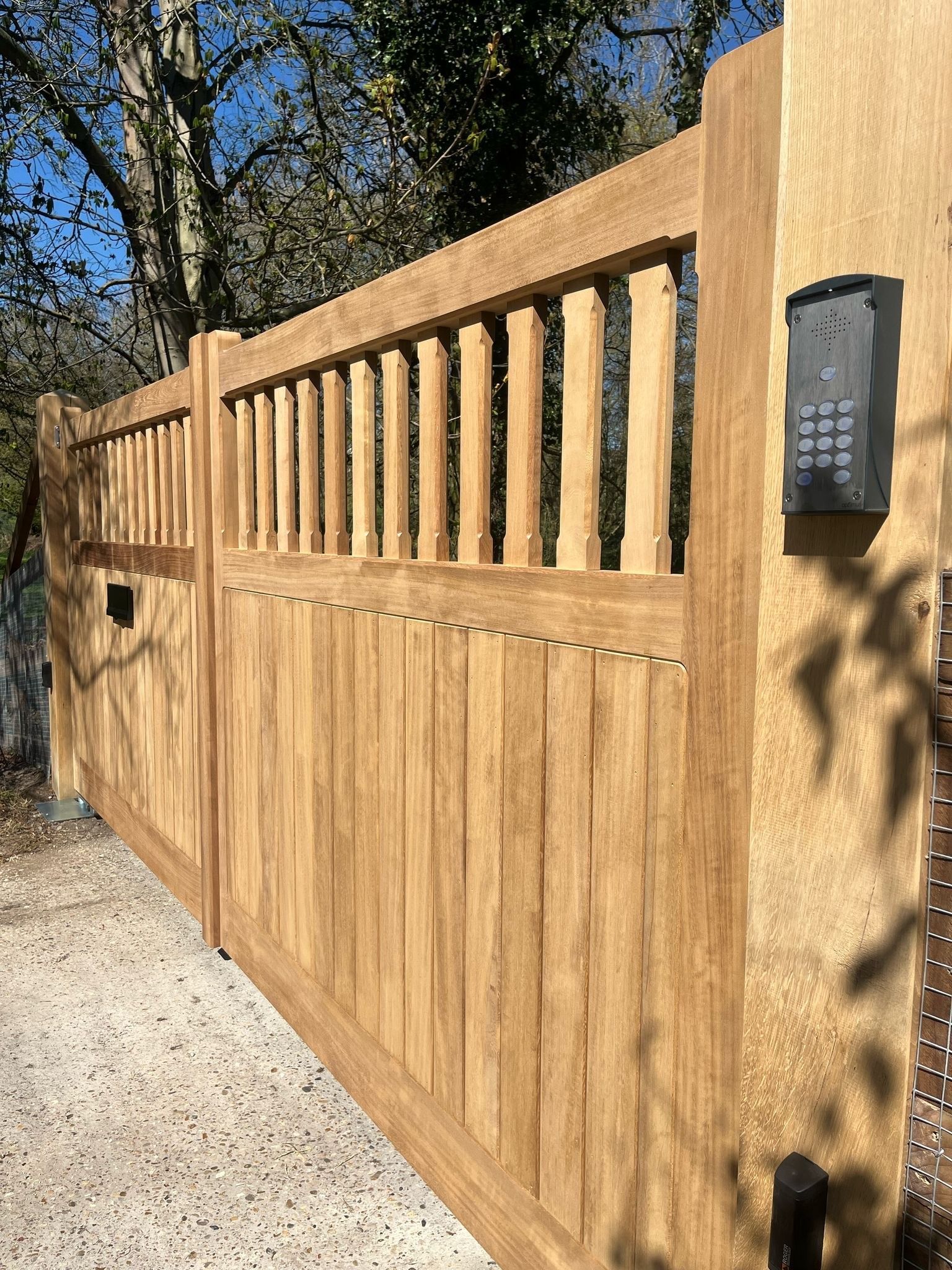 Wooden privacy gate with vertical slats and keypad entry, set beside a gravel driveway and trees.