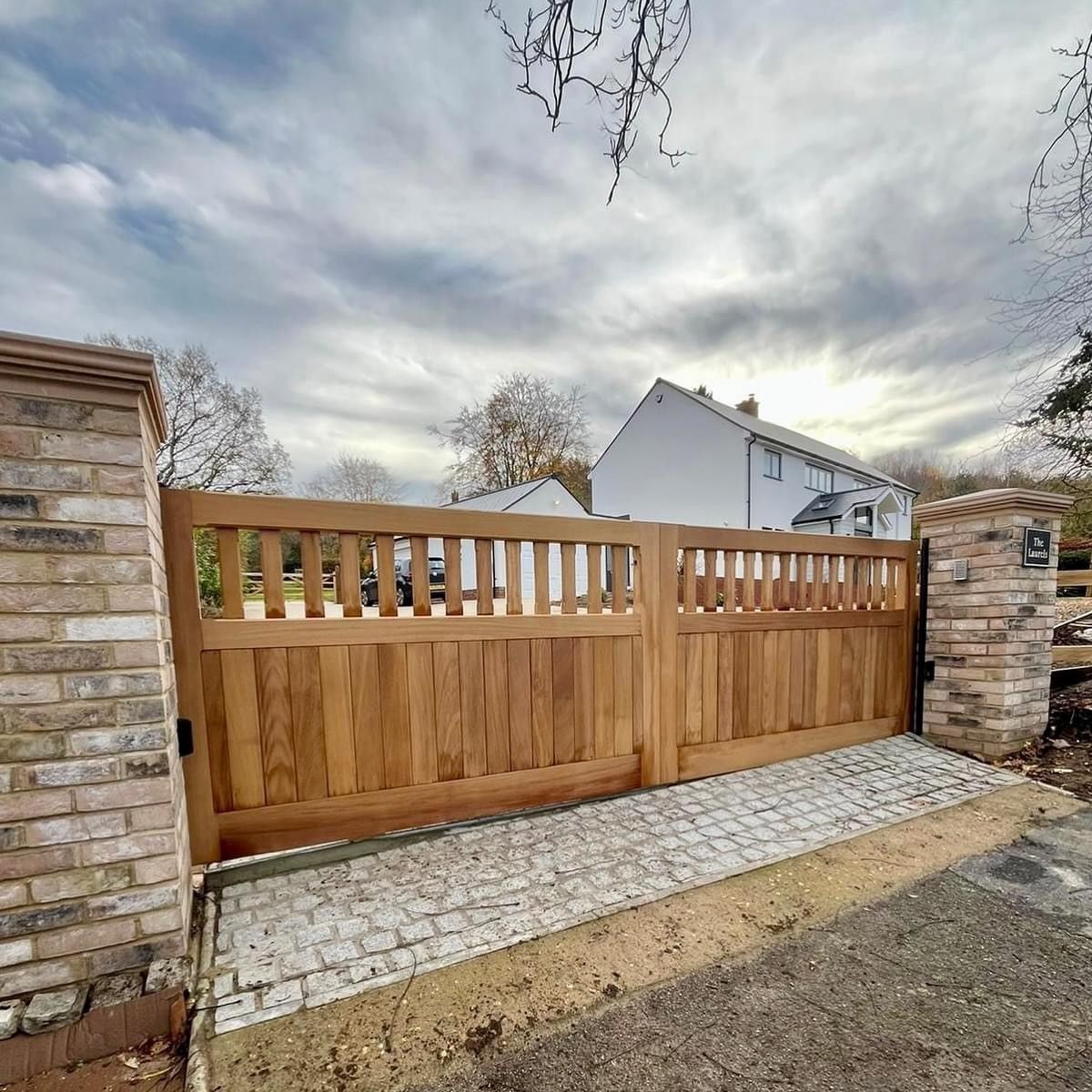 Wooden driveway gate between brick pillars beside a cobblestone path under a cloudy sky