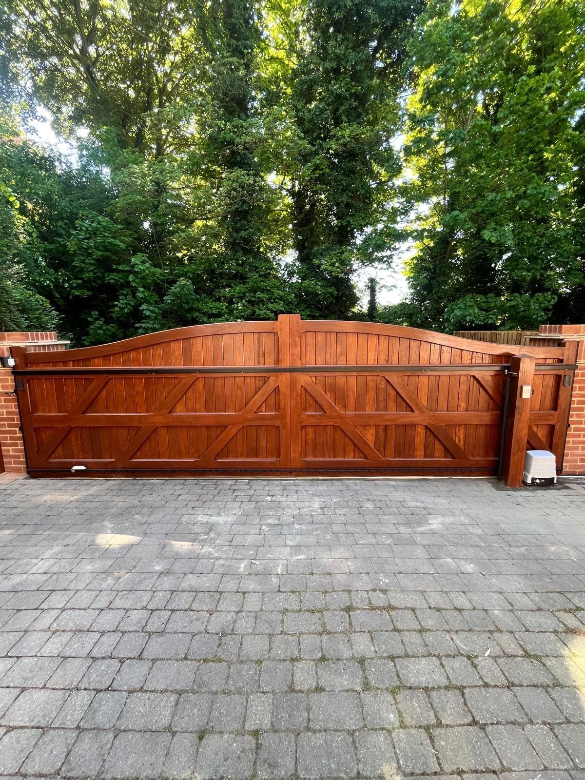 Wooden driveway gate with brick pillars, set behind a paved courtyard and tall green trees.