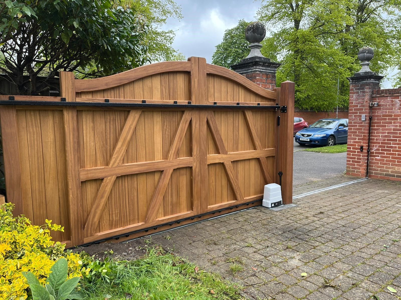 Wooden driveway electric gate beside a brick wall, with a cobblestone driveway and parked cars beyond.