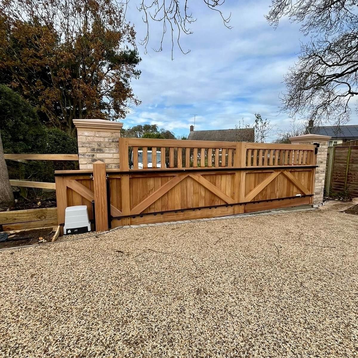 Wooden driveway electric gate with stone pillars and gravel foreground under a cloudy sky