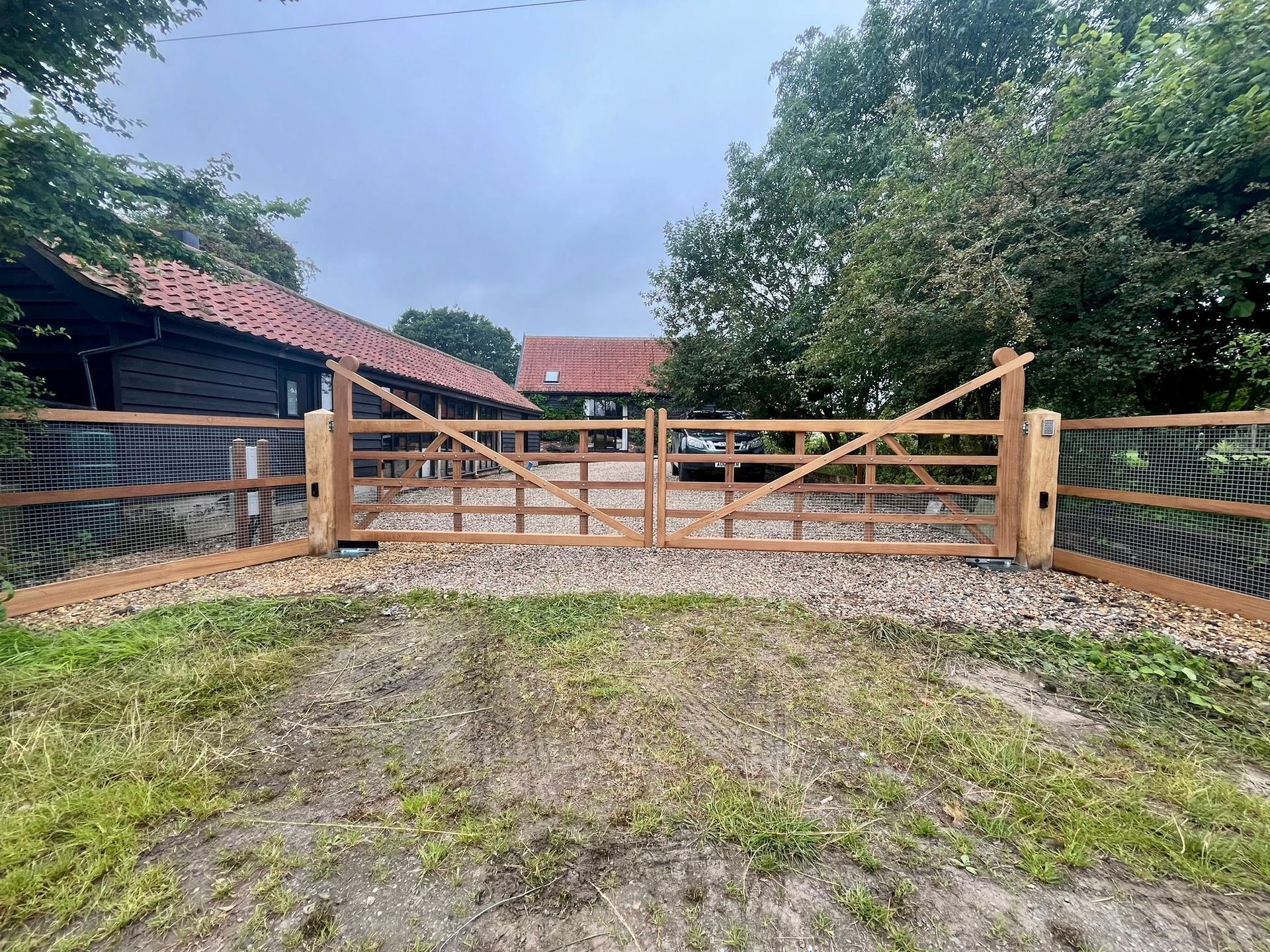 Wooden farm electric gate and fence at a gravel driveway, with red-roofed buildings and trees in the background