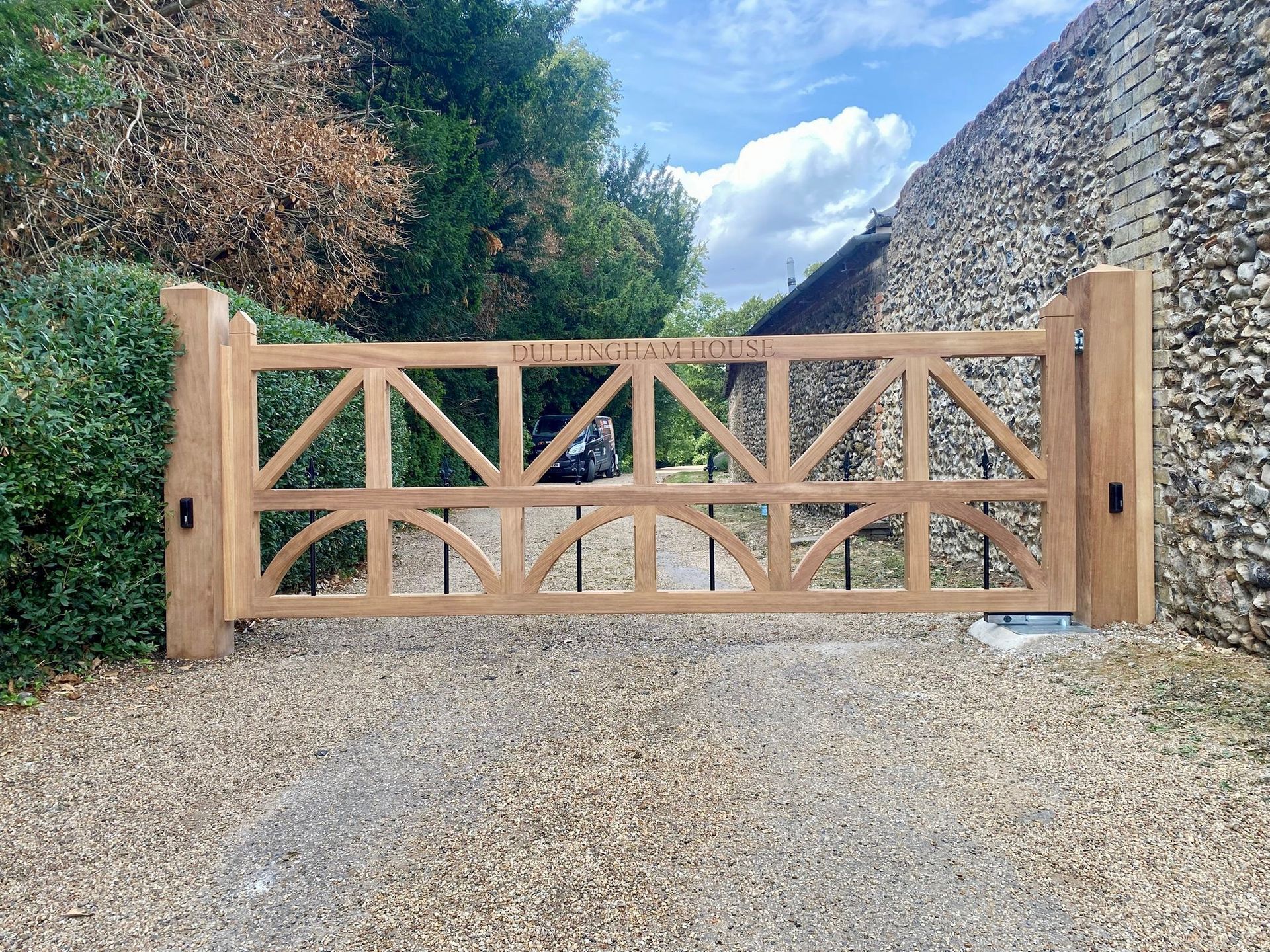Wooden driveway electric gate across a gravel entrance between hedges and a stone wall