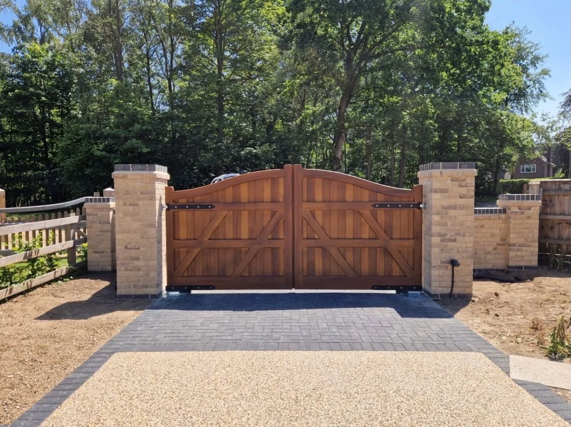 Wooden driveway gate between stone pillars, with trees and a gravel path in front.