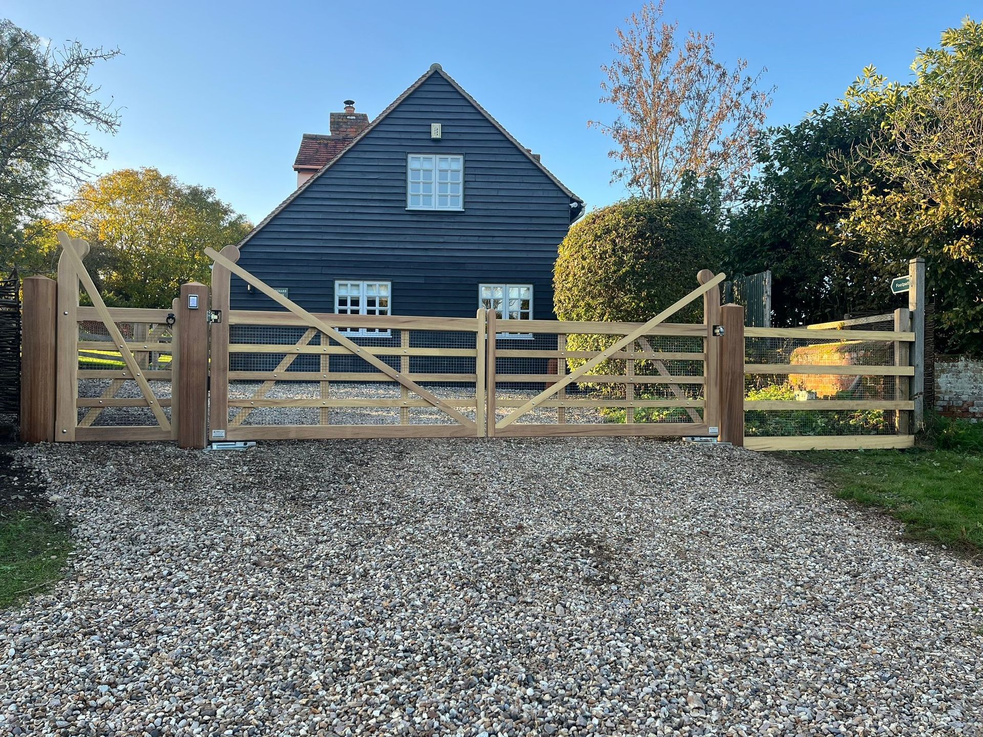 Wooden electric gate and fence before a dark blue house with gravel driveway and trees in front