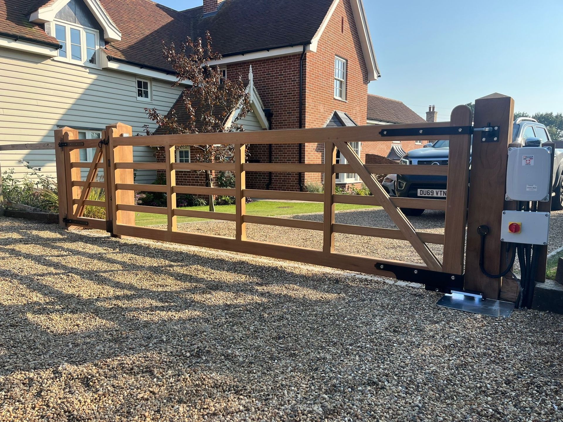 Wooden driveway electric gate beside brick houses, with gravel foreground and gate motor post on the right.
