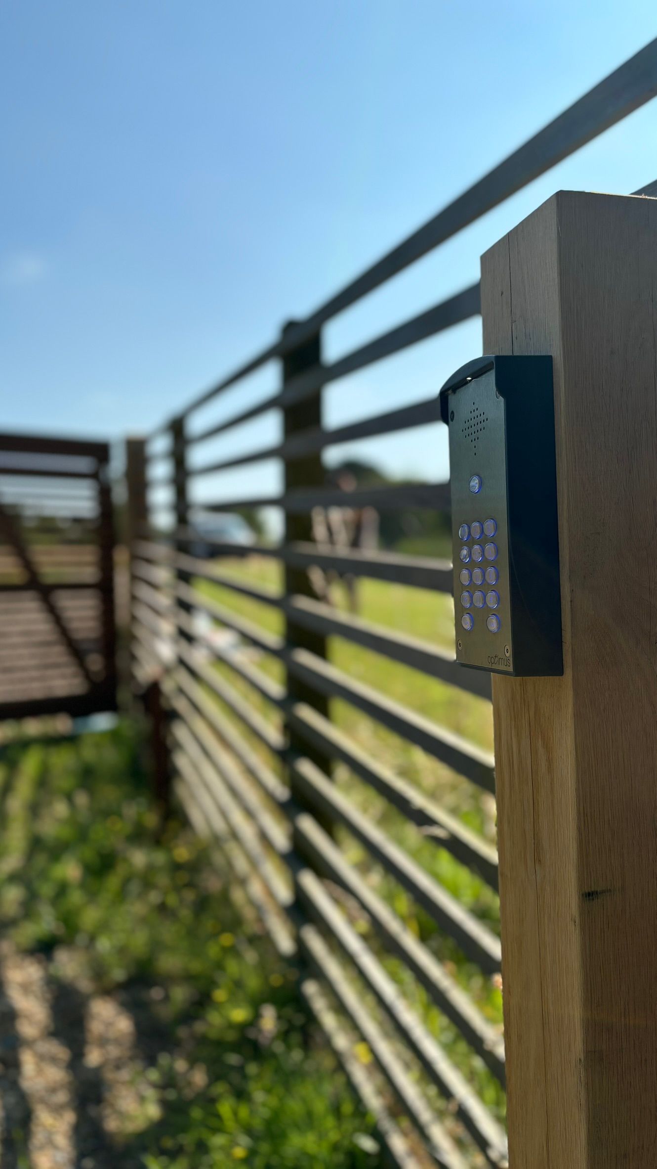 Close-up of a wooden fence post with a dark gate latch, sunlit field and metal fence in the background