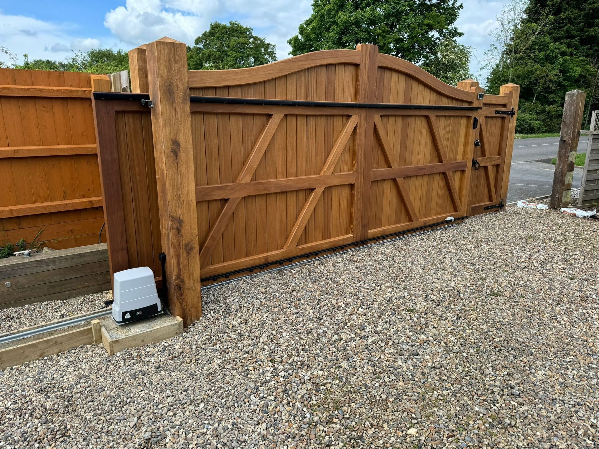 Wooden driveway gate with diagonal bracing beside a gravel path and fence, outdoors under a cloudy sky
