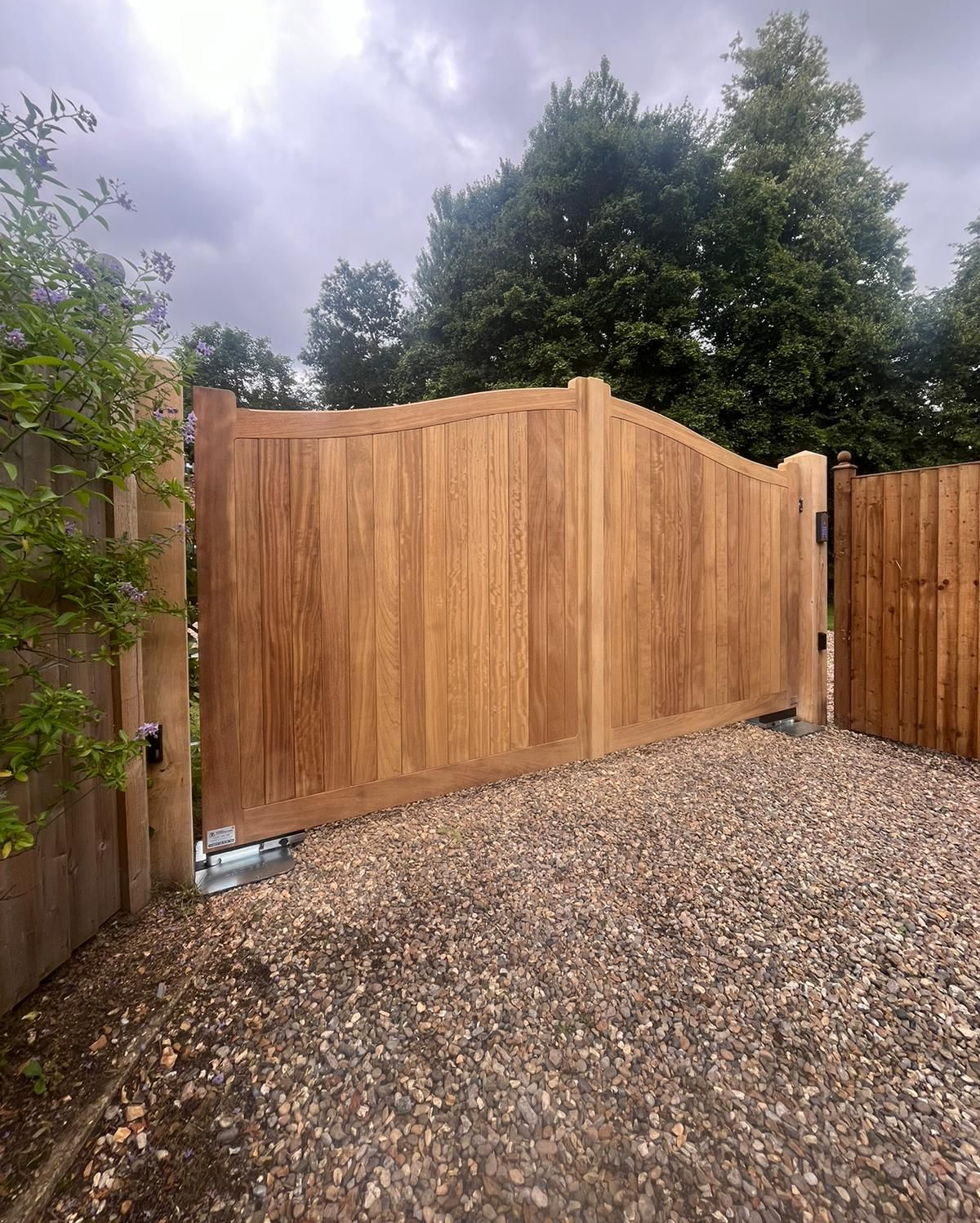 Wooden privacy fence and electric gate beside a gravel driveway under cloudy skies.