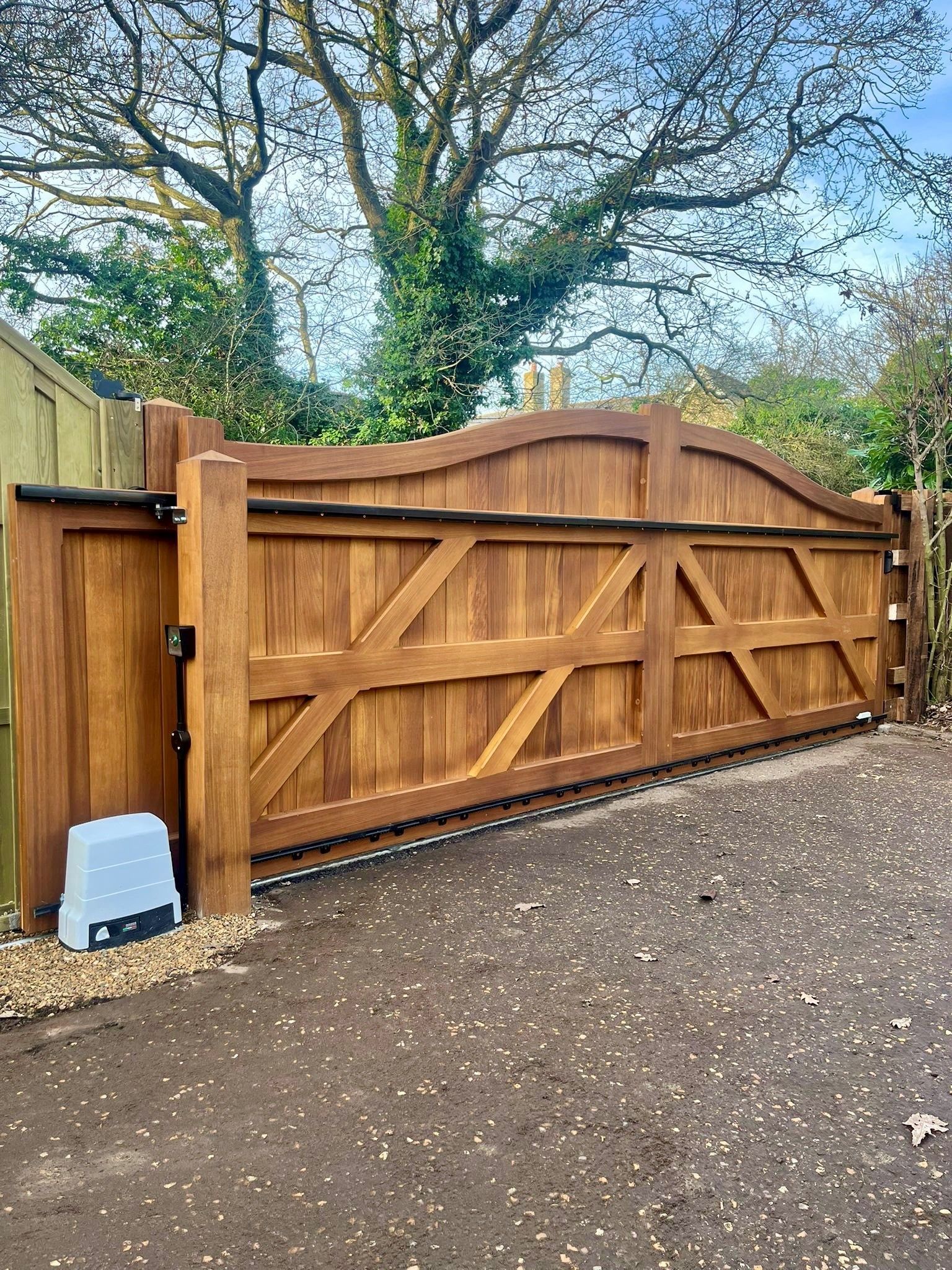 Wooden driveway gate with diagonal braces and a side post, set beside a gravel path and trees.