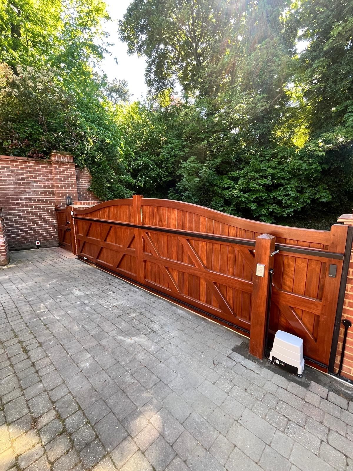 Wooden driveway gate beside a brick wall, opening onto a paved yard with dense greenery behind.