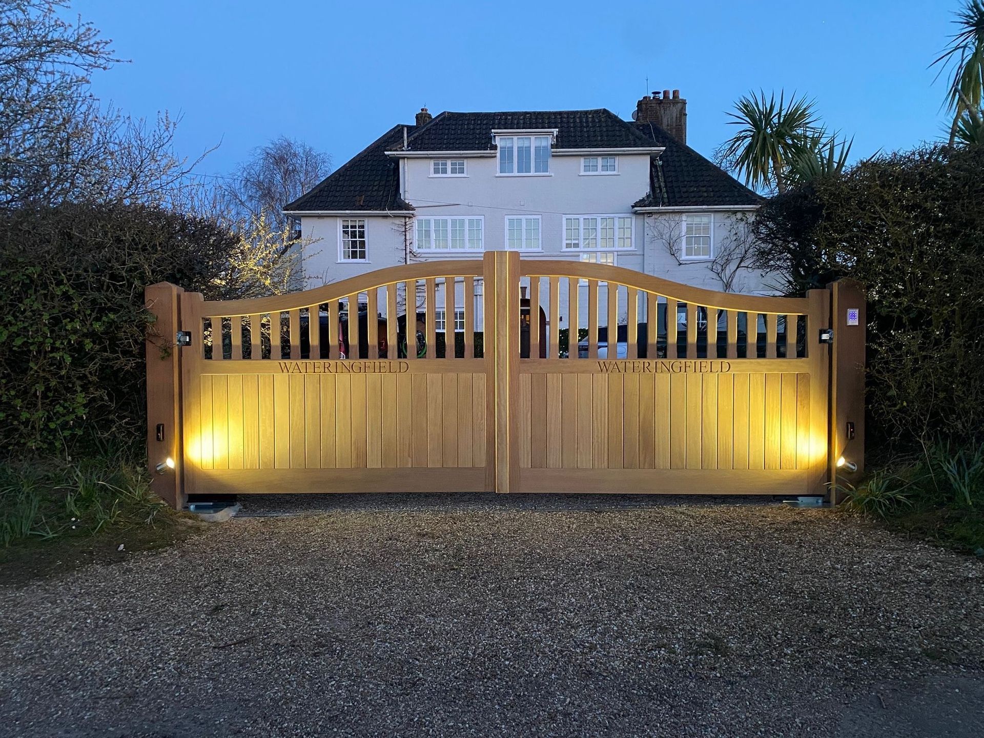 Illuminated wooden driveway electric gate at dusk in front of a large white house