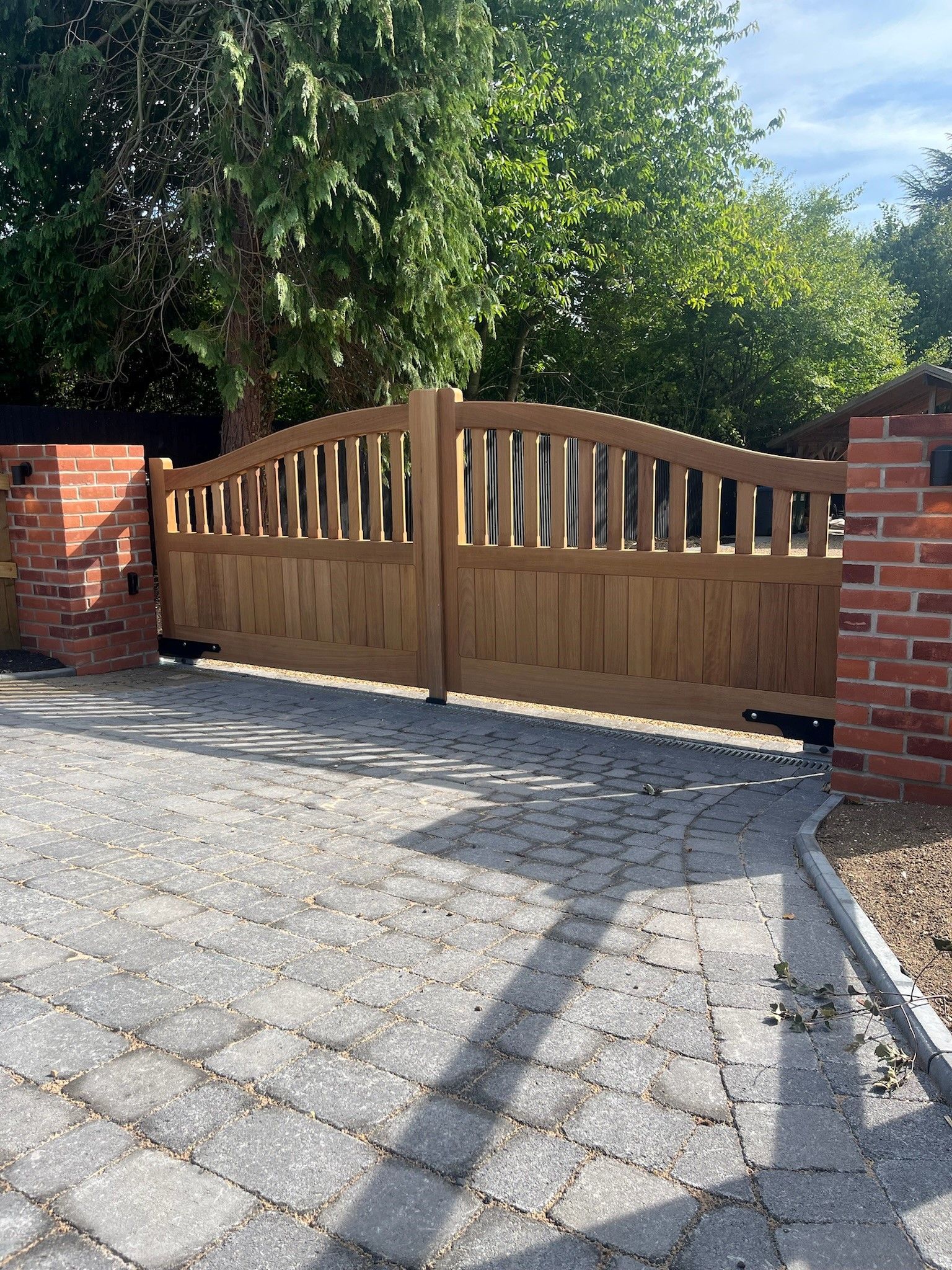 Wooden driveway electric gate between red brick pillars with gravel foreground and leafy trees behind