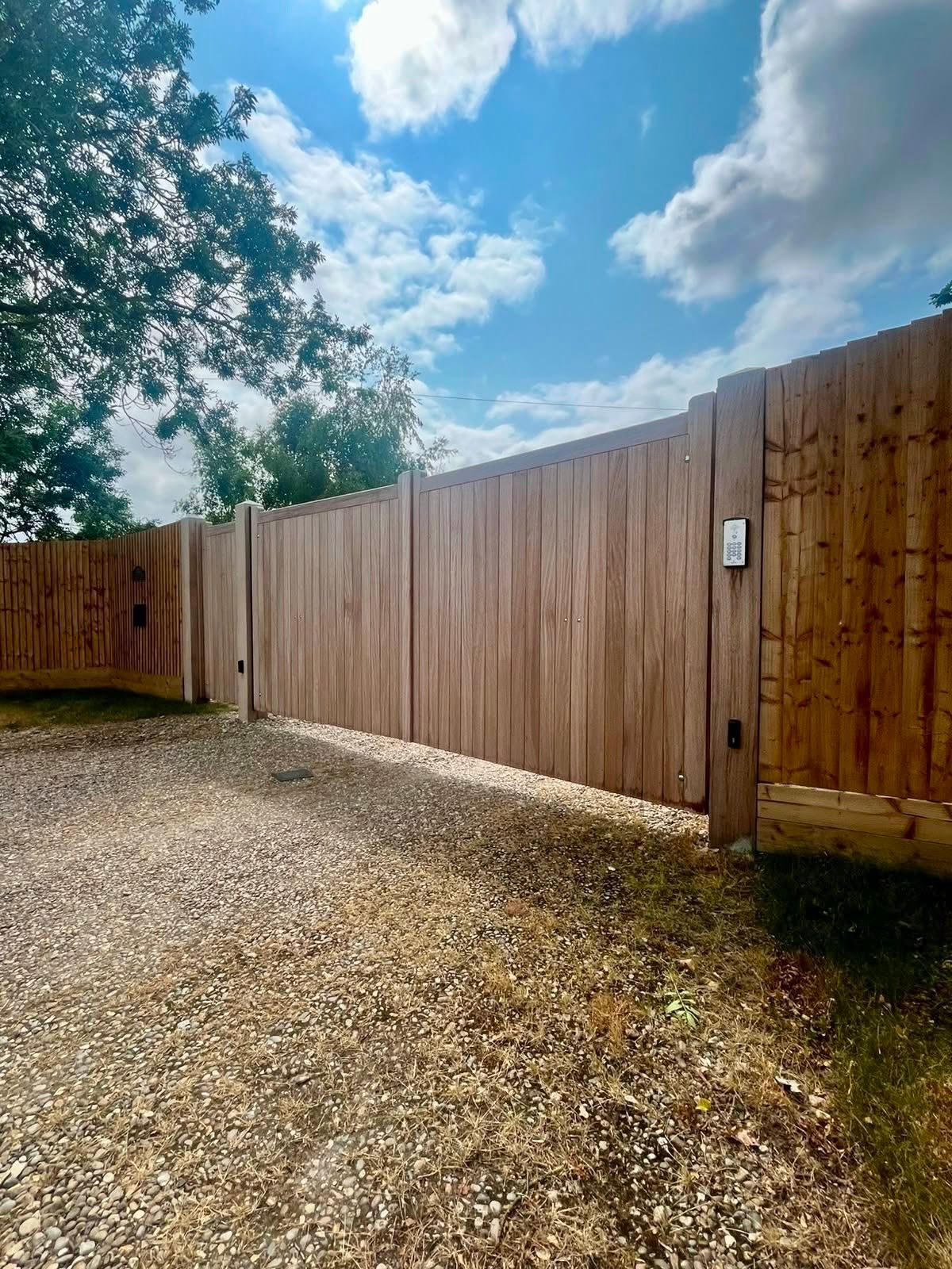 Gravel driveway beside a tall wooden fence under a partly cloudy blue sky