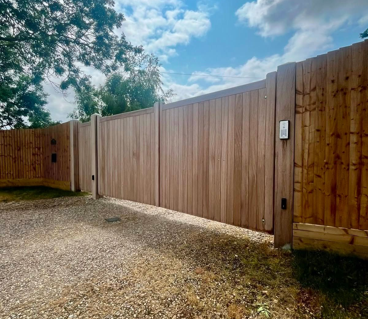 Gravel driveway beside a tall wooden privacy fence under a partly cloudy sky