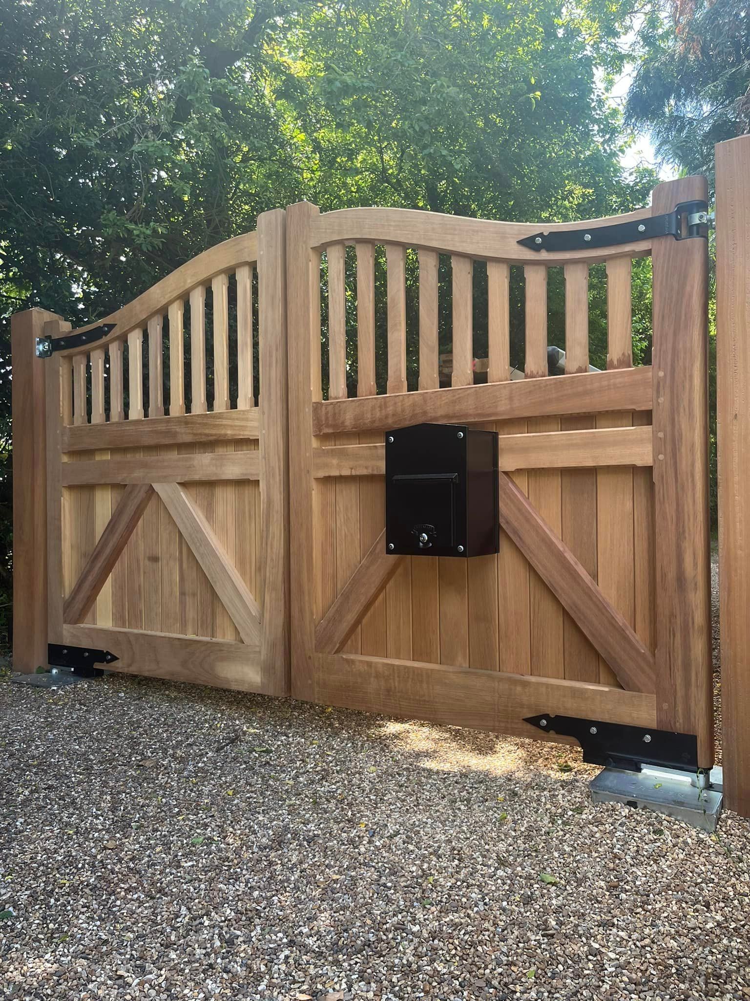 Wooden driveway electric gate with black mailbox, gravel foreground, and green trees behind.