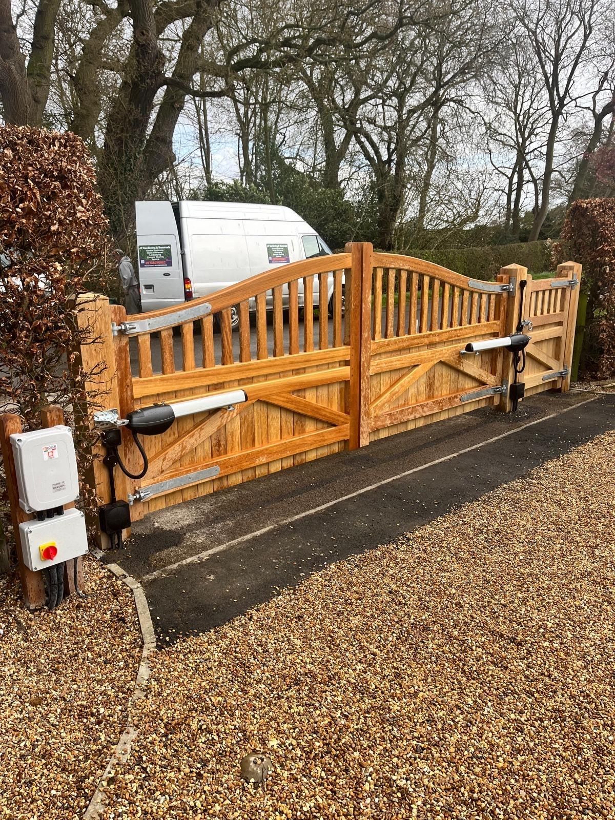 Wooden driveway electric gate with black motorized posts beside a gravel path and parked white van