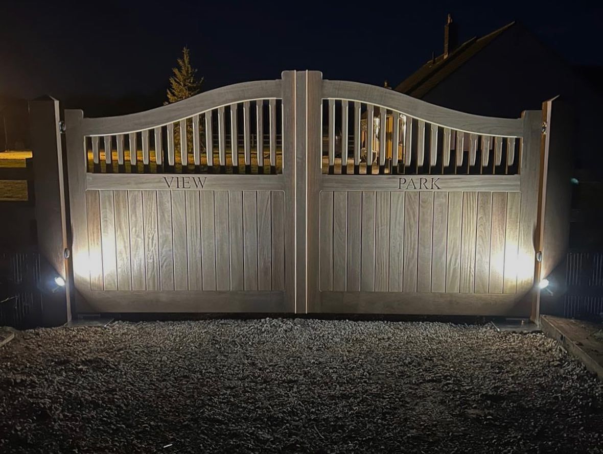 Illuminated white metal electric gate at night with curved slats and gravel driveway in front
