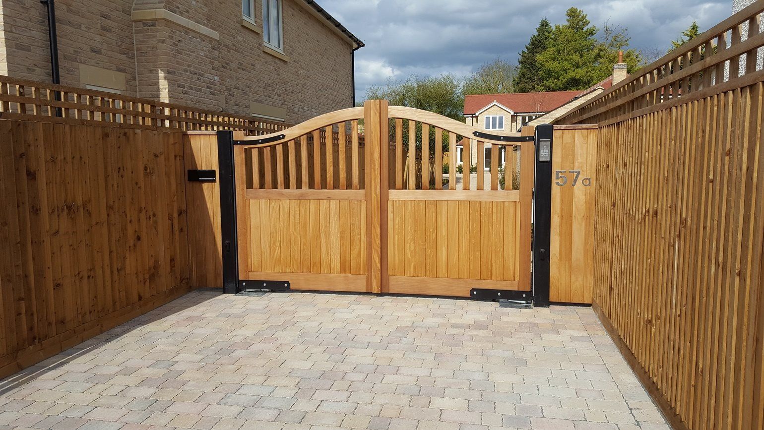 Stone driveway leading to a closed wooden electric gate between tall wooden fences under a cloudy sky