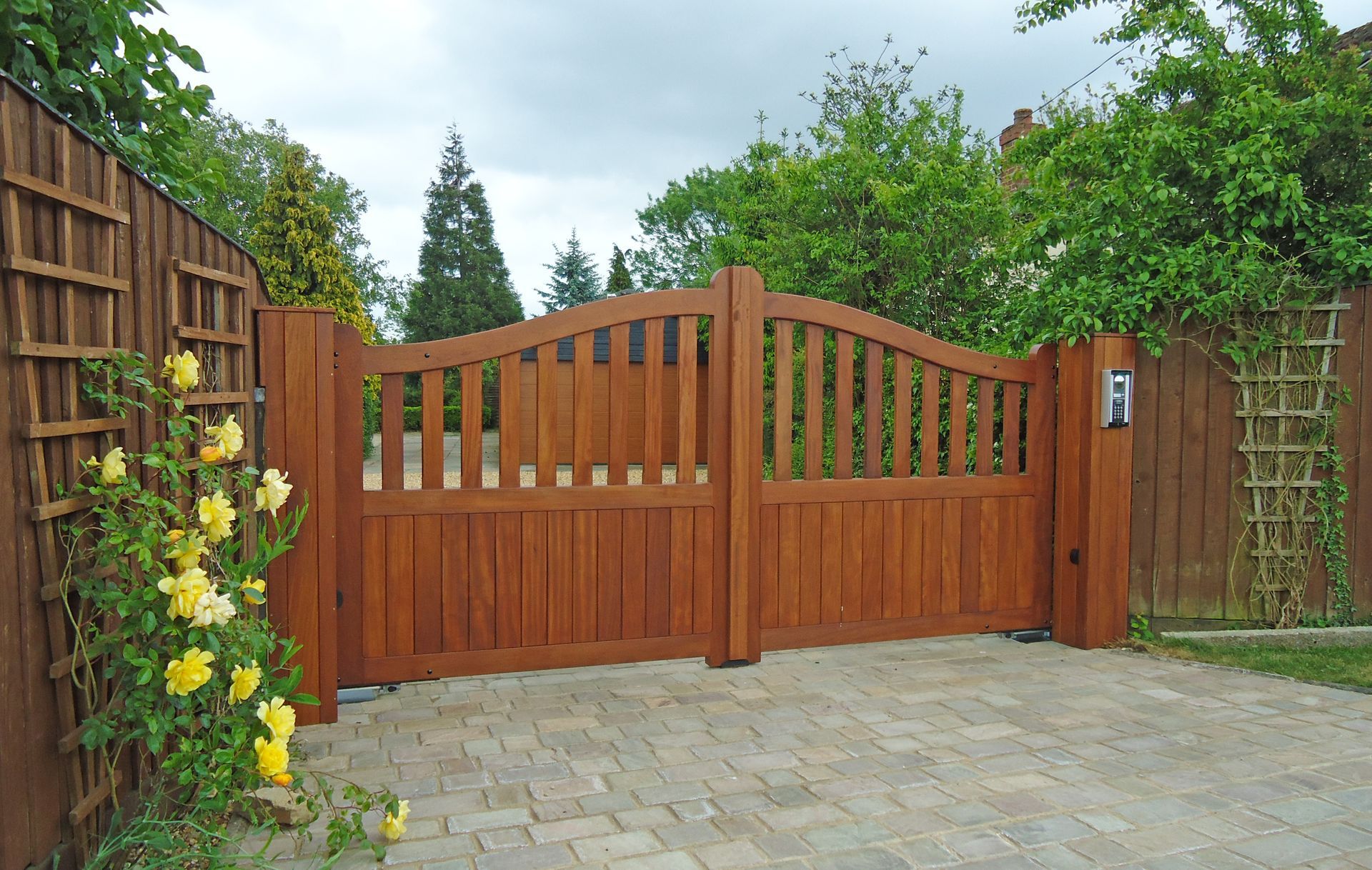 Wooden double electric gate with arched slats in a stone driveway, framed by green hedges and vines.