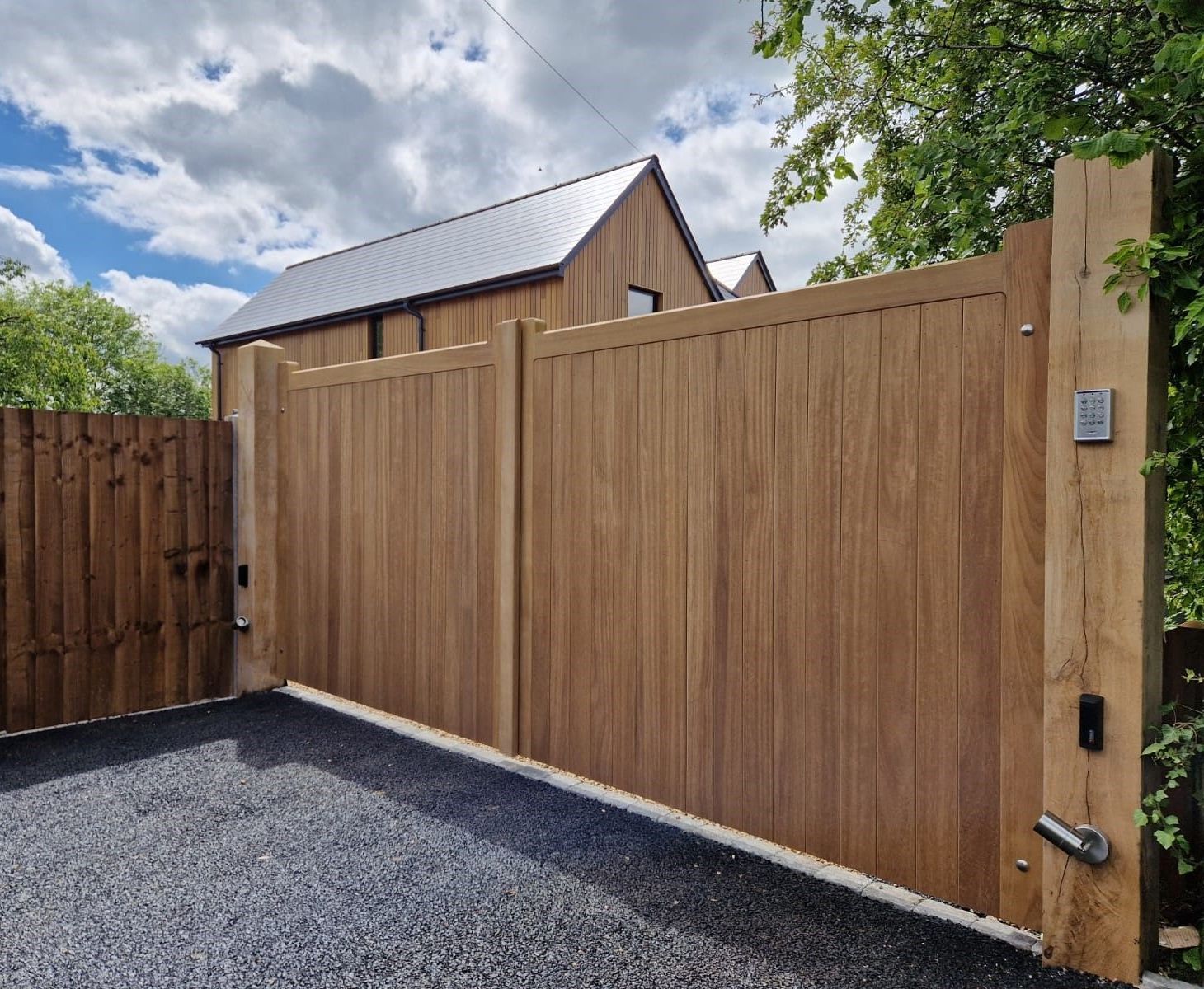 Modern wooden driveway gate and fence beside a gravel yard under a cloudy sky