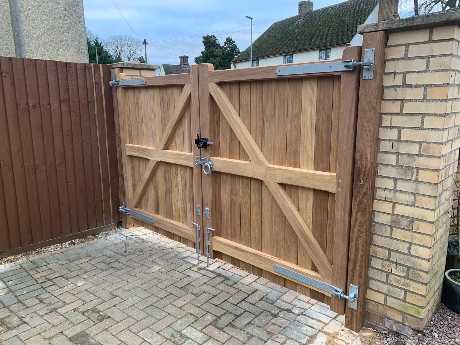 Wooden driveway gate with diagonal braces and metal hinges beside a brick wall and paved courtyard