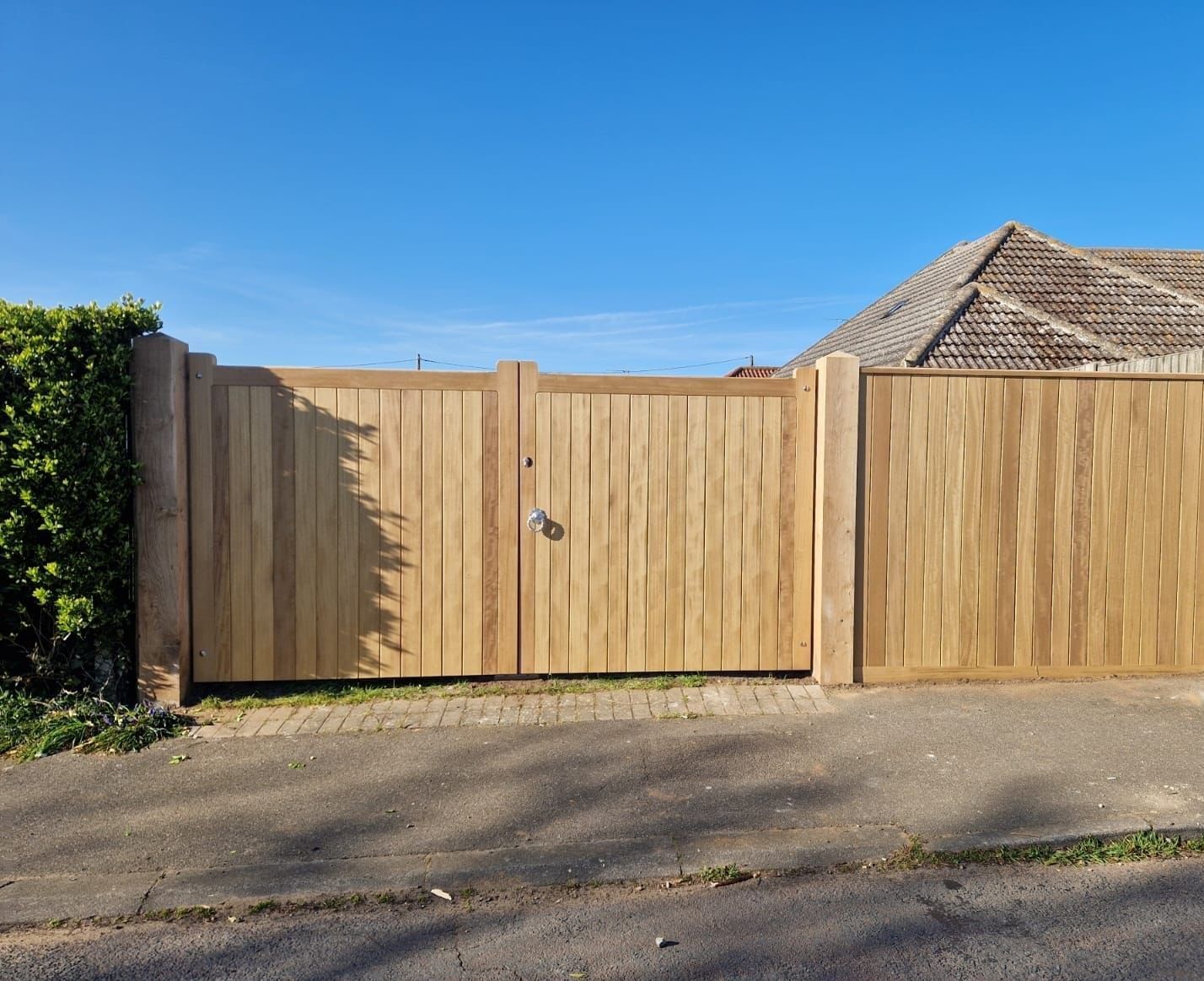 Wooden driveway gate beside a hedge and brick wall under a clear blue sky.