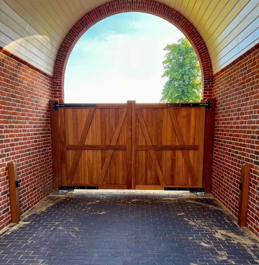 Wooden gate set in a brick archway with a paved driveway beneath a bright sky.