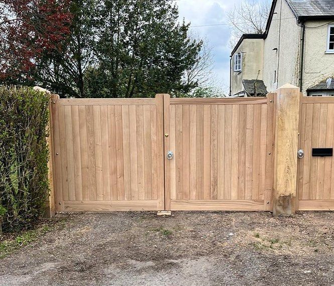Wooden driveway gate with a central latch, set beside a hedge and stone house.