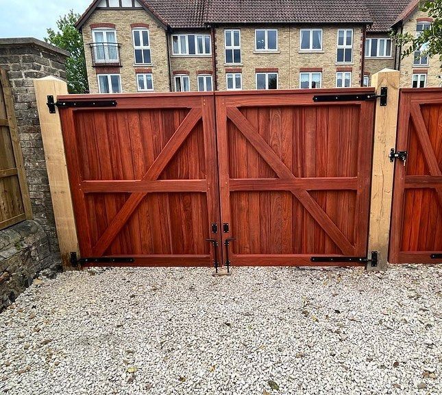 Double wooden driveway gates with black hinges in front of a brick house, beside a gravel path