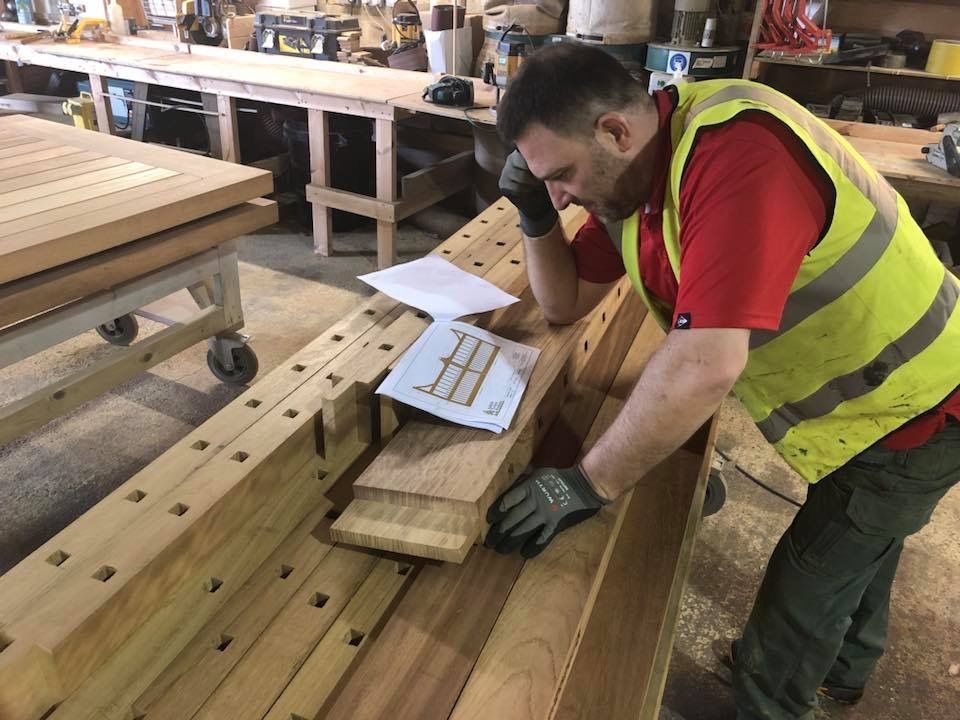 Worker in a yellow safety vest measuring a wooden board at a workbench in a workshop.