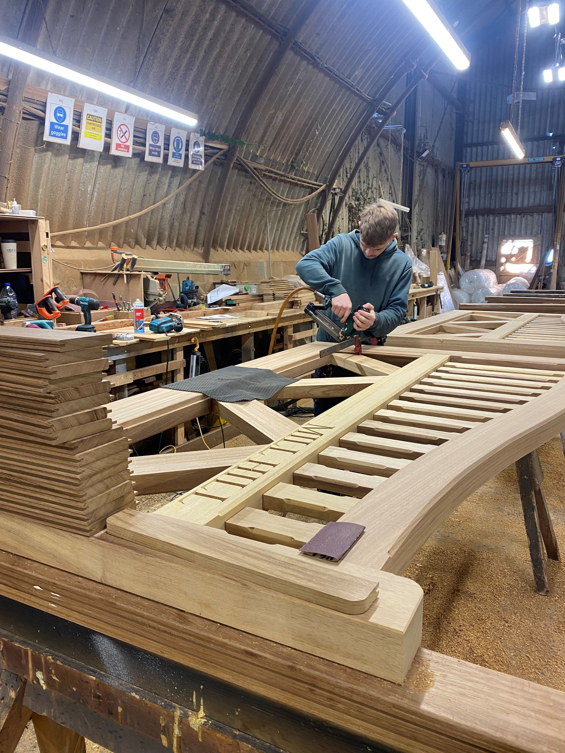 Person sanding a large wooden boat hull in a workshop with tools and lights overhead