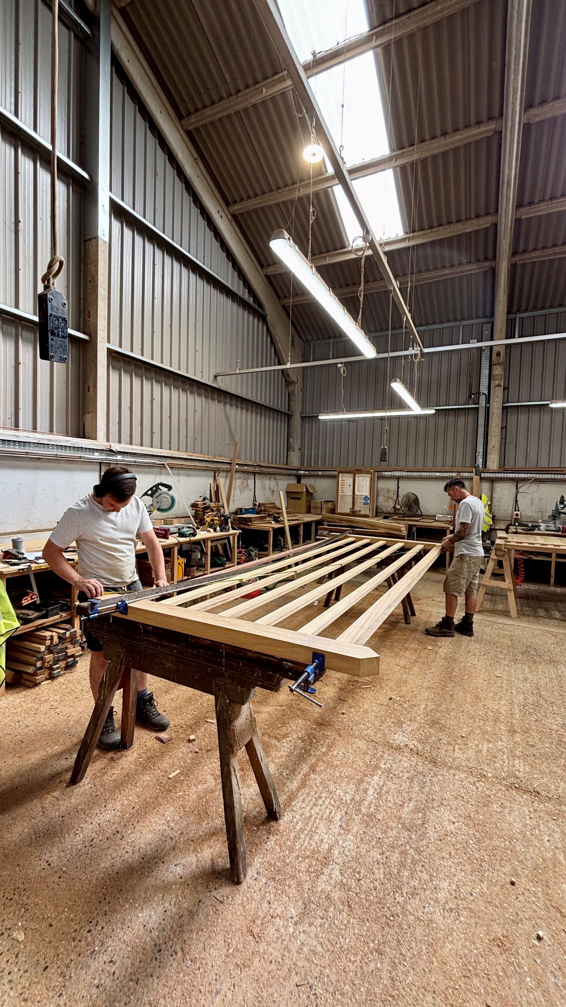 Workers assembling long wooden planks inside a bright workshop with sawdust-covered floor.