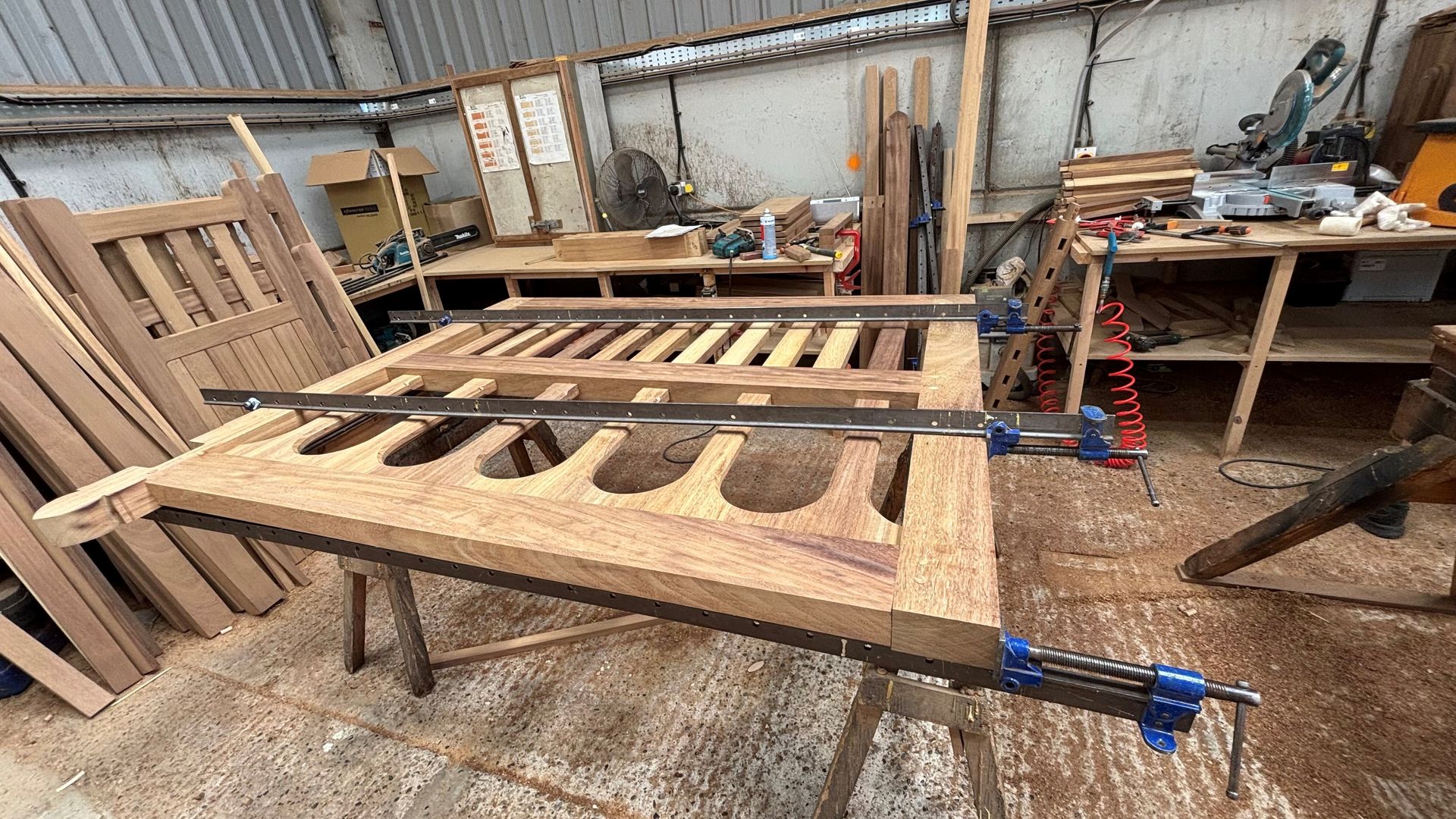 Wooden panel clamped on a workbench in a rustic workshop, with cutout openings and tools nearby.