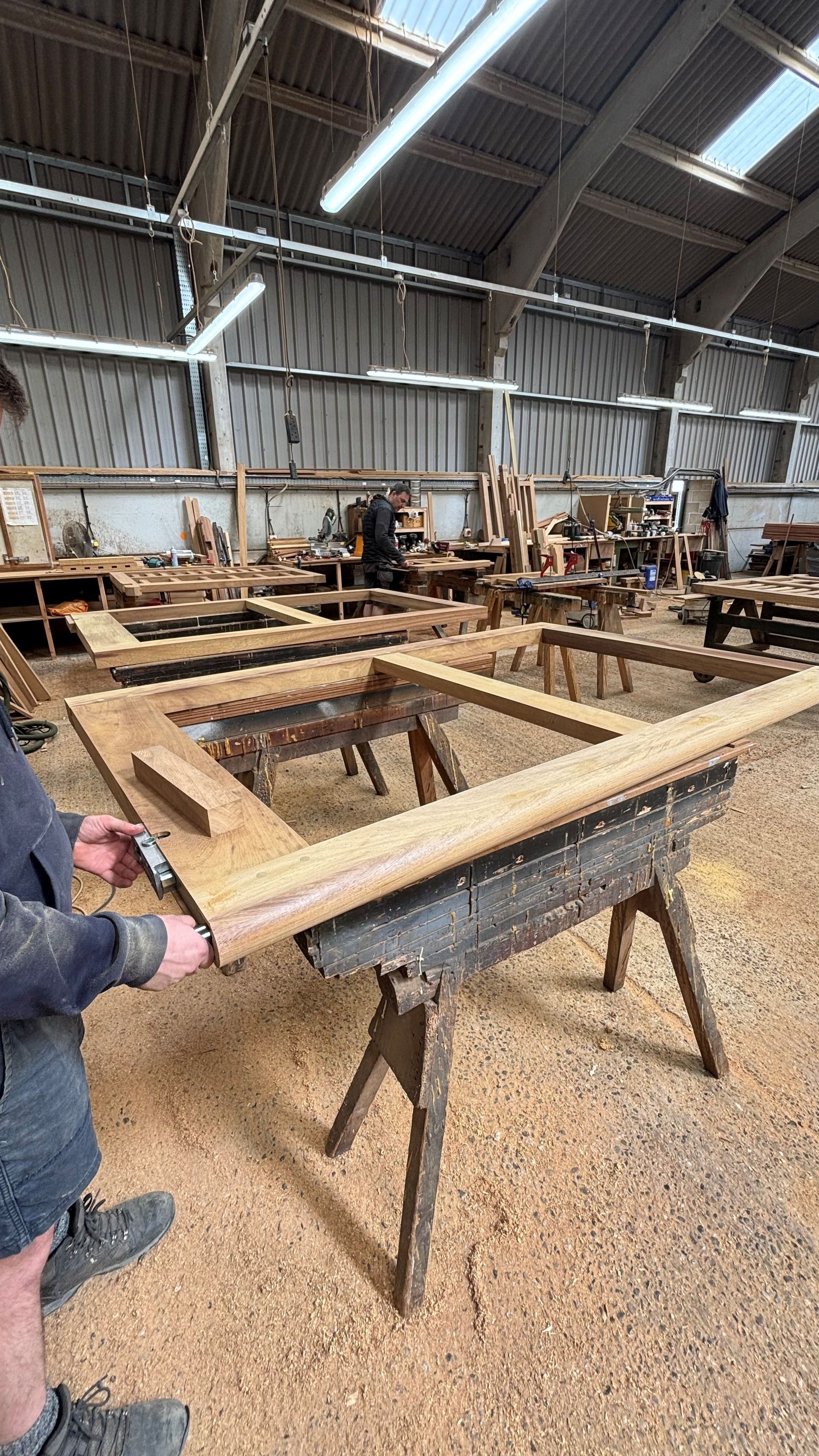 Workshop with large wooden frames on sawhorses; a person in a jacket holds one frame in the foreground.