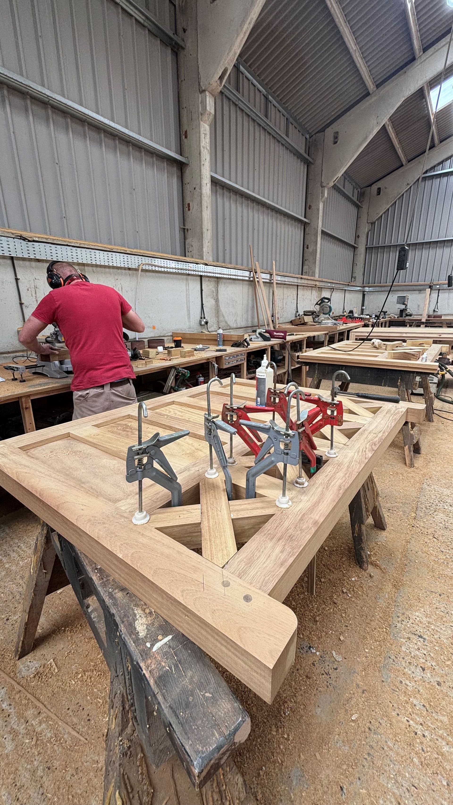 Worker in red shirt assembling wooden frames inside a large workshop with tools and sawdust.